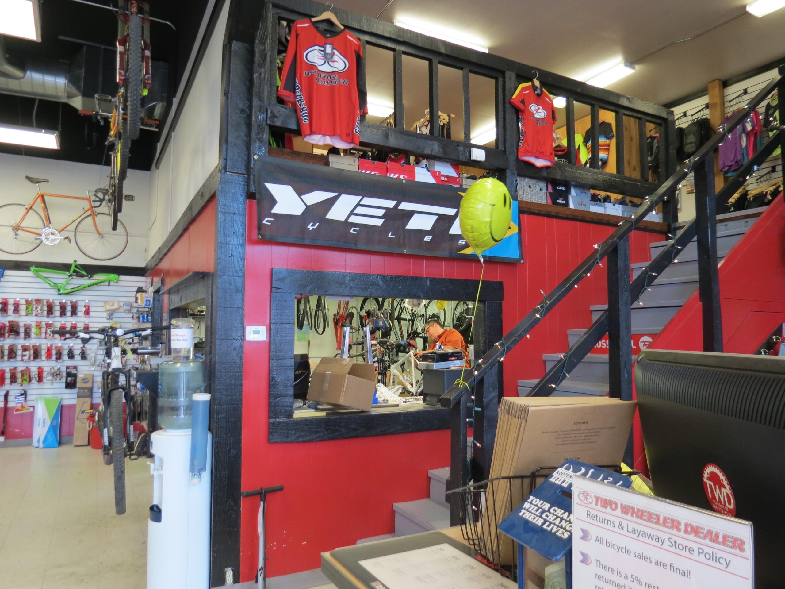 Interior view of a bicycle shop featuring a red and black color scheme. The store includes a raised area with cycling jerseys hanging on display. A bicycle repair area is visible in the background, while the main counter showcases various bike-related items and a water cooler. A yellow smiley balloon adds a touch of color to the scene.