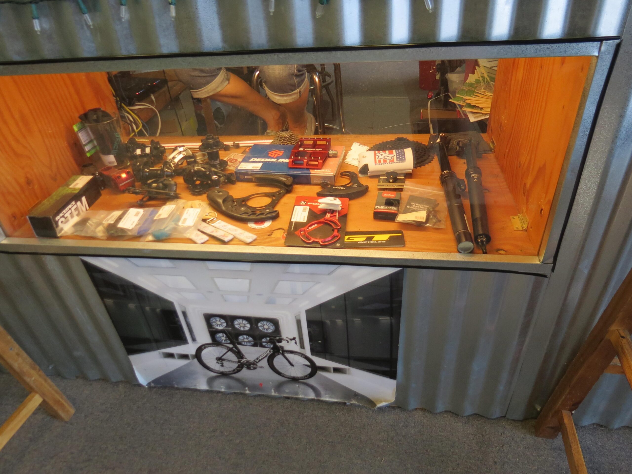 A display case containing various bicycle parts and accessories, including tools, gears, and components, with a cyclist graphic on the bottom panel. The background shows a partial view of a person sitting behind the case.