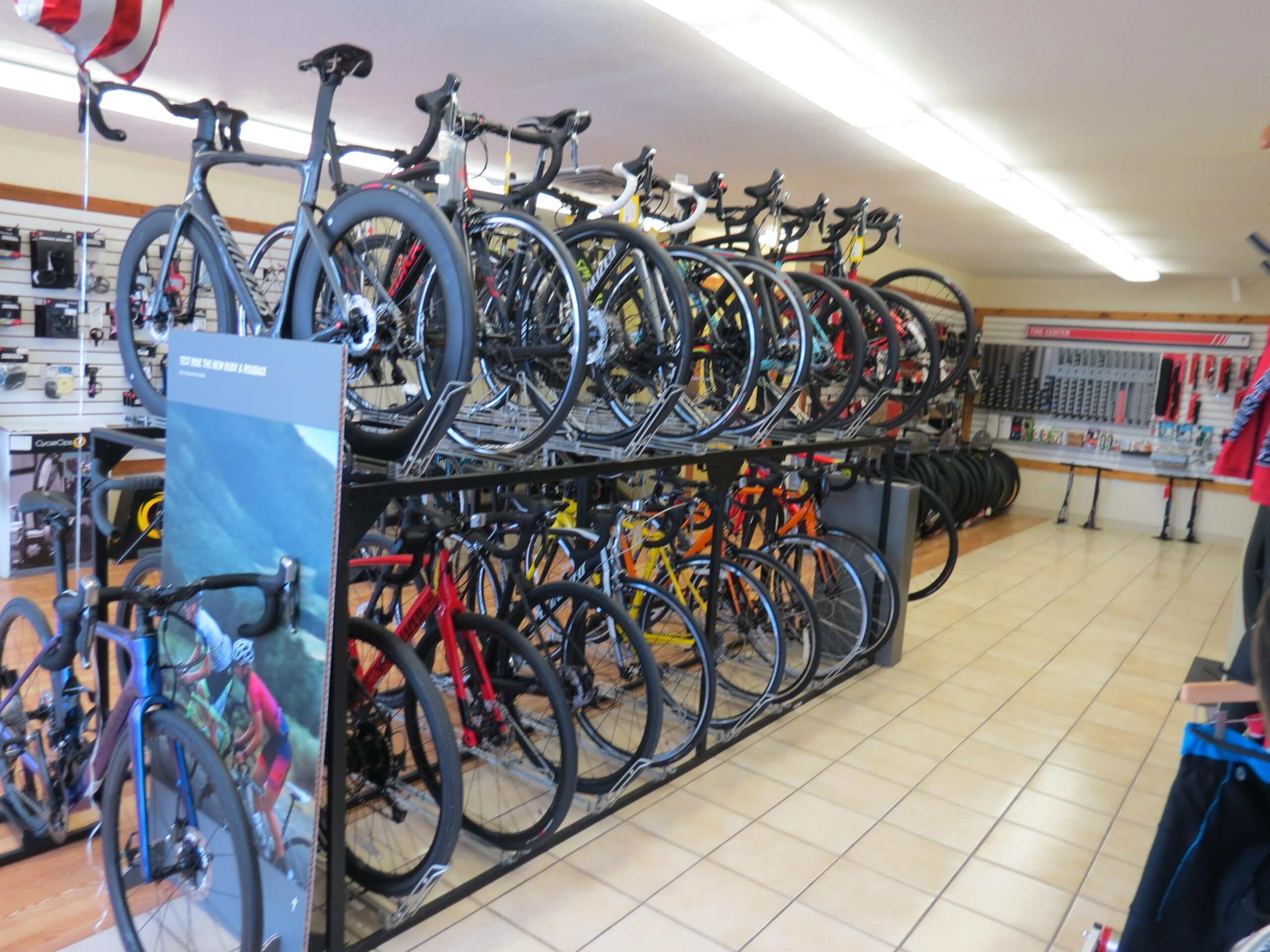 A bicycle shop interior featuring multiple rows of bicycles displayed on racks, including a variety of colors and styles. The shop has a tiled floor and shelves in the background displaying cycling accessories and equipment. Soft lighting illuminates the space.