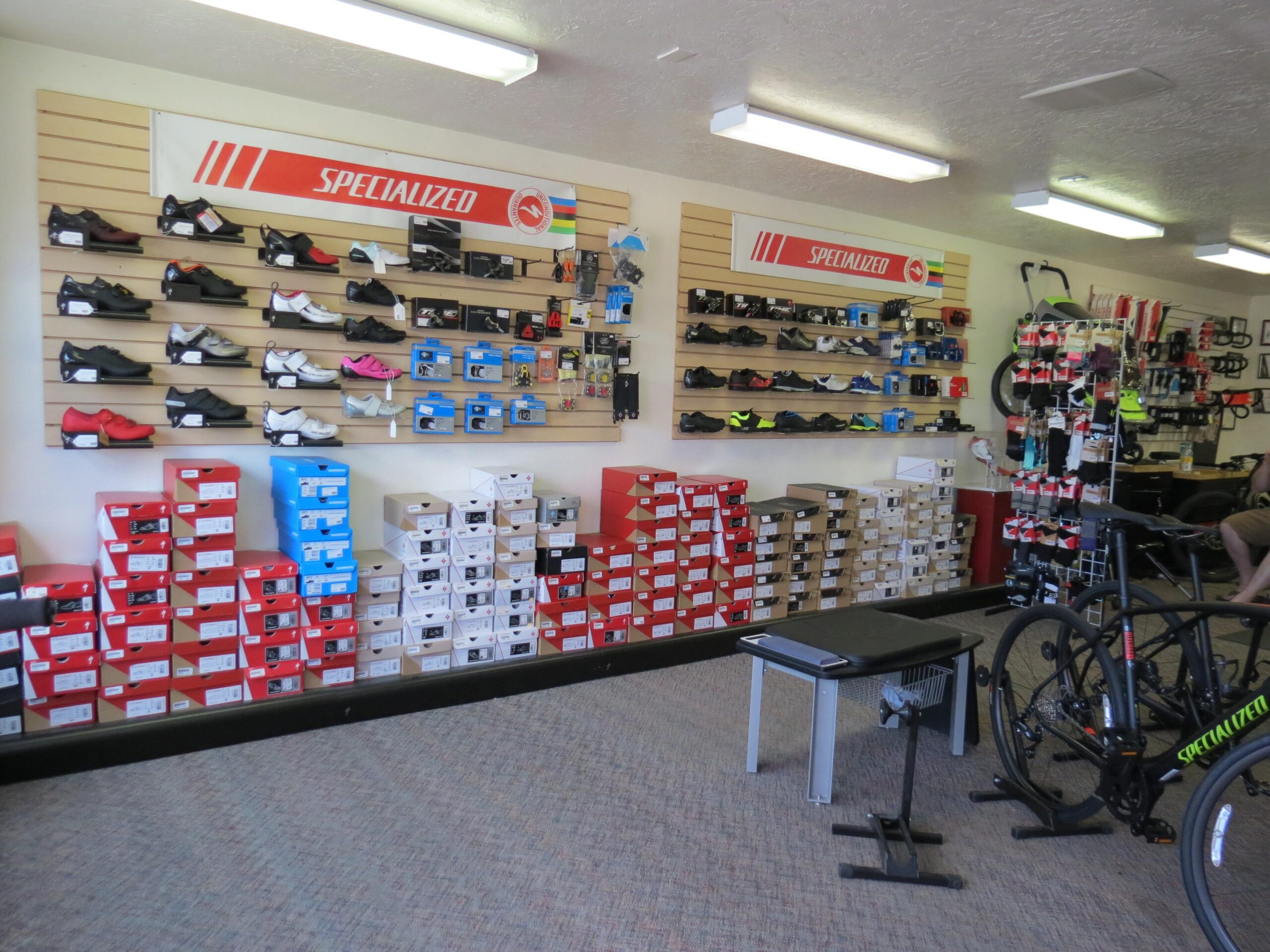 Interior view of a bicycle retail store featuring a wall display of cycling shoes, a variety of shoe boxes stacked on the floor, and biking accessories. A bike stand with a bicycle is visible in the foreground, and shelves hold additional gear. The overall setup reflects a well-organized space dedicated to cycling enthusiasts.