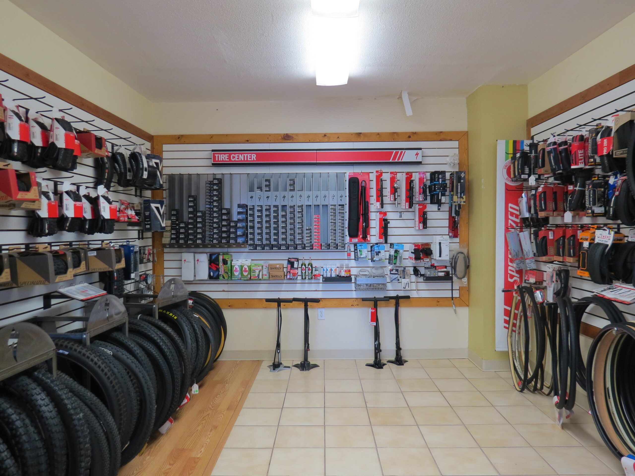 Interior of a tire center shop displaying an array of bike tires and accessories. The walls are lined with visible products, including tires, tools, and maintenance items, organized on pegboards. There are two workstations in the center of the room, with tile flooring and bright overhead lighting.