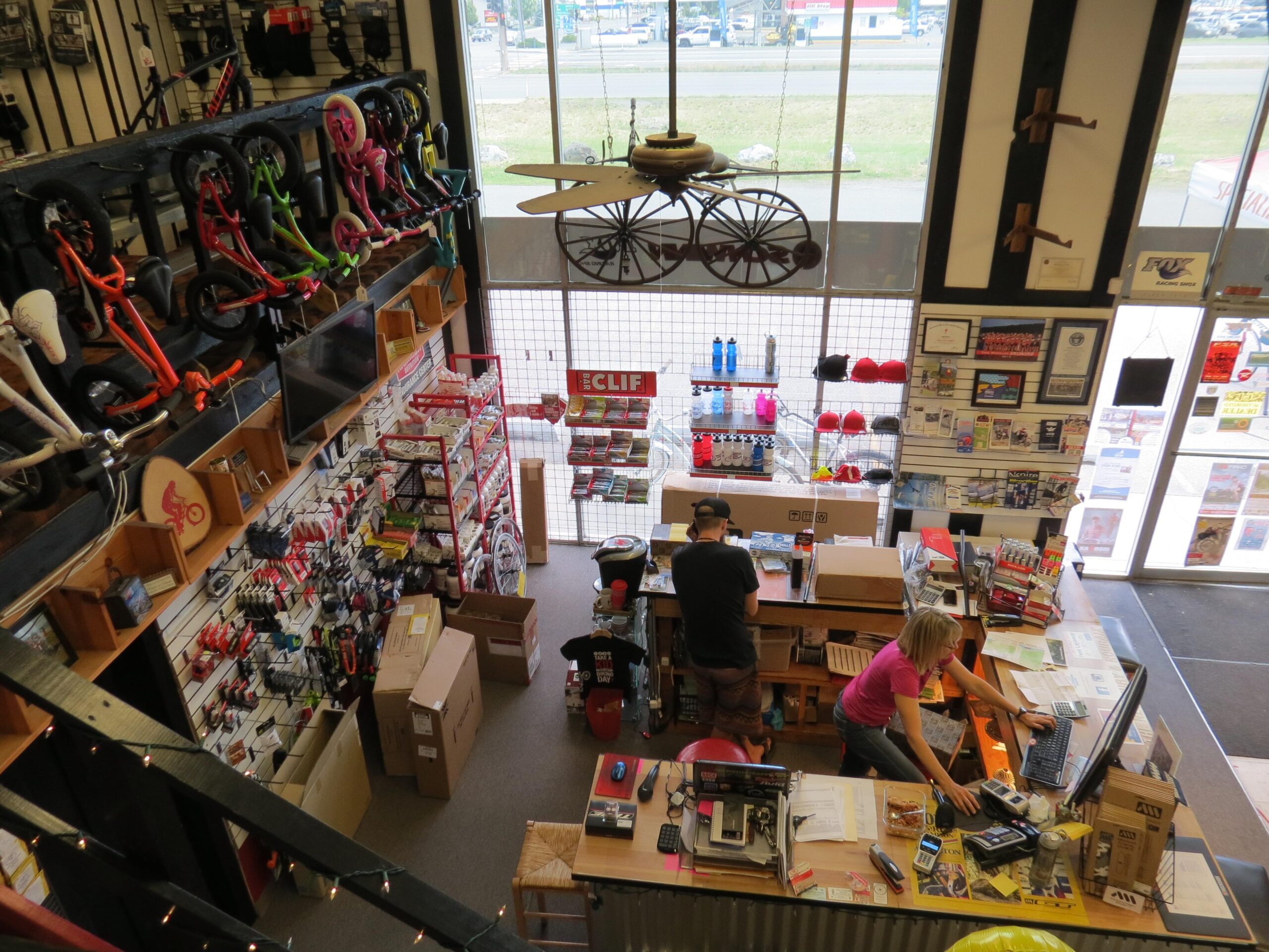 A view from above inside a retail store showcasing a variety of bicycles displayed on the walls. Two employees are engaged in tasks at the checkout counter, with one using a computer and the other working with packages. The store features shelves filled with sporting goods and merchandise, while large windows provide natural light and a view of the street outside.