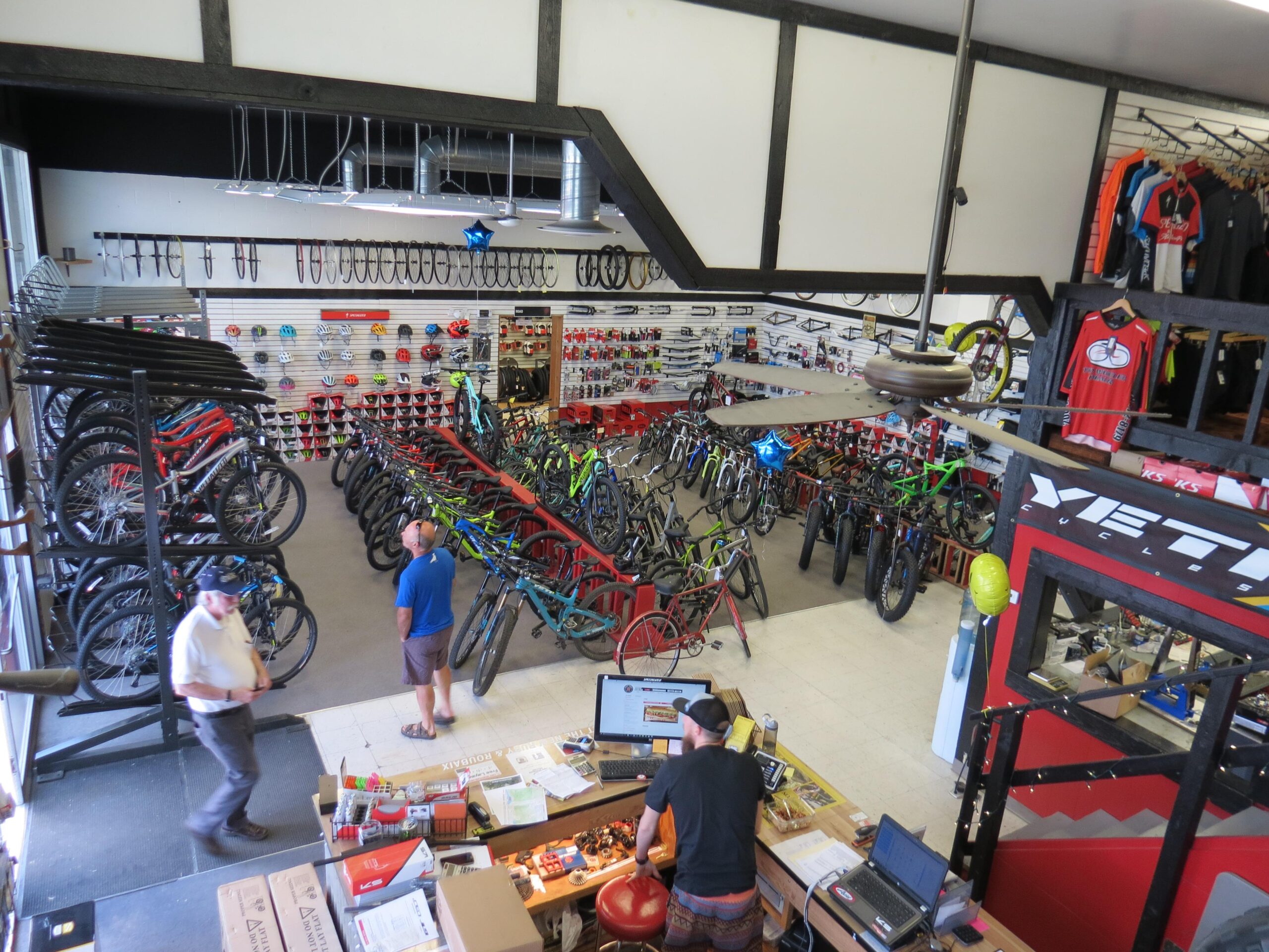 A view of a bicycle shop showcasing a wide selection of bikes organized on racks, with various helmets and accessories displayed on the walls. Two customers are browsing the bikes, while a staff member is at the sales counter working on a computer. The shop has a bright interior with a fan hanging from the ceiling and is decorated with cycling apparel.