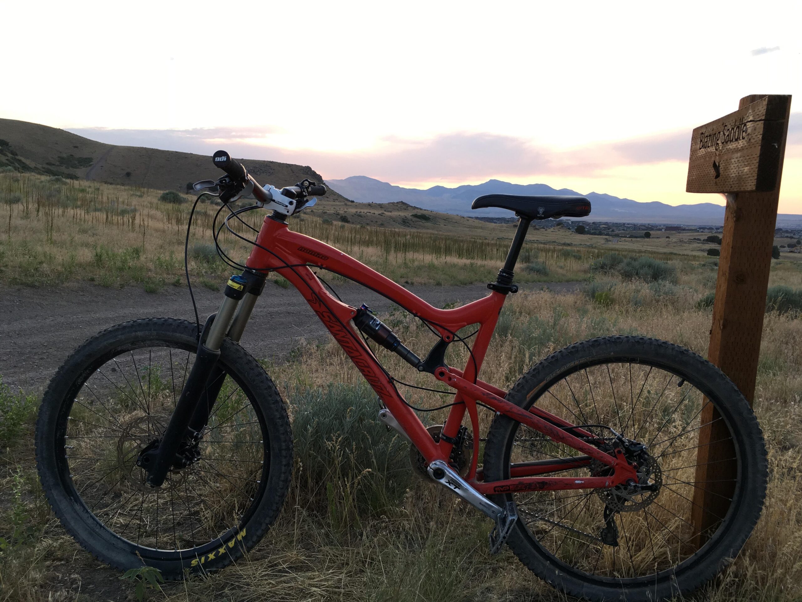 Santa Cruz Nomad: A red mountain bike leaning against a wooden sign that reads "Blazing Saddle," set against a backdrop of rolling hills and a colorful sunset. Tall grass and sparse vegetation frame the scene, emphasizing the outdoor setting.