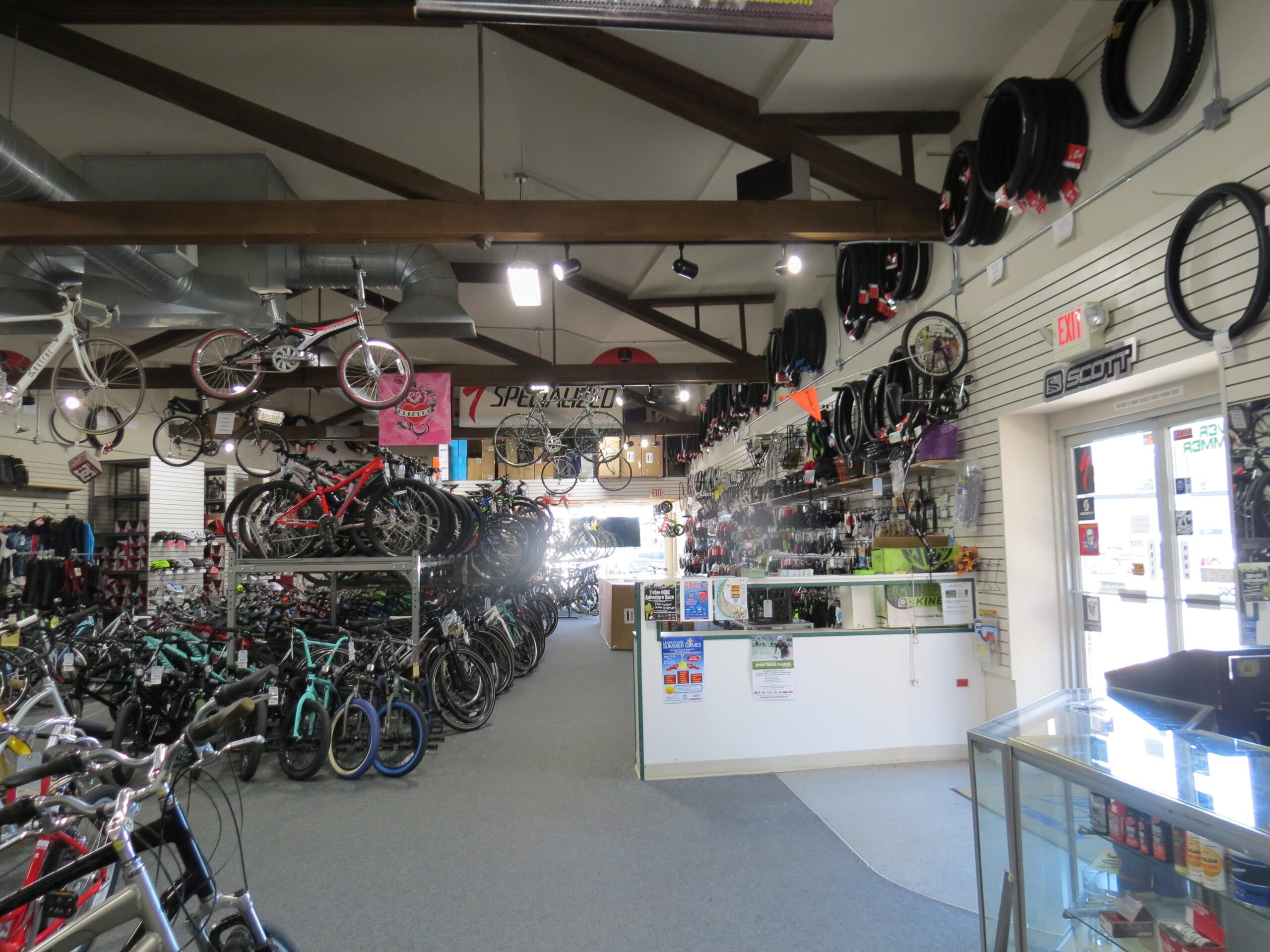 Interior view of a bicycle shop featuring a variety of bicycles displayed on the floor and hanging from the ceiling. Bike accessories and tires are mounted on the walls, and a service area is visible at the front with a glass display case. The shop has a bright and welcoming atmosphere, highlighted by wooden beams in the ceiling.