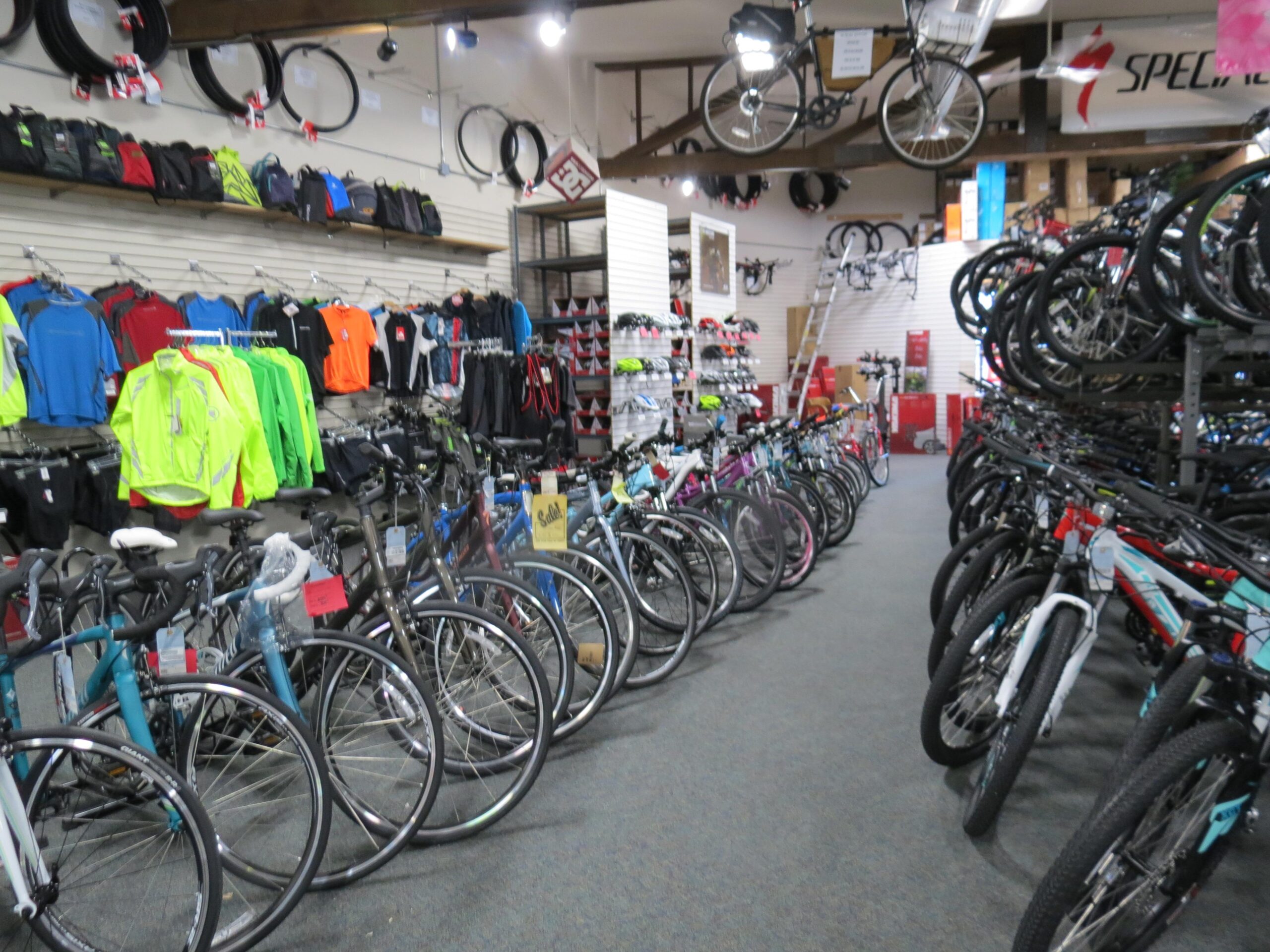 A well-organized bicycle shop interior featuring a wide selection of bicycles along one side, with various styles and colors on display. The opposite wall displays cycling apparel, including brightly colored jerseys and jackets, along with accessories like backpacks. Overhead, bike tires are neatly arranged, and additional bicycles are hung from the ceiling. The store has a tidy layout, allowing customers to browse comfortably.