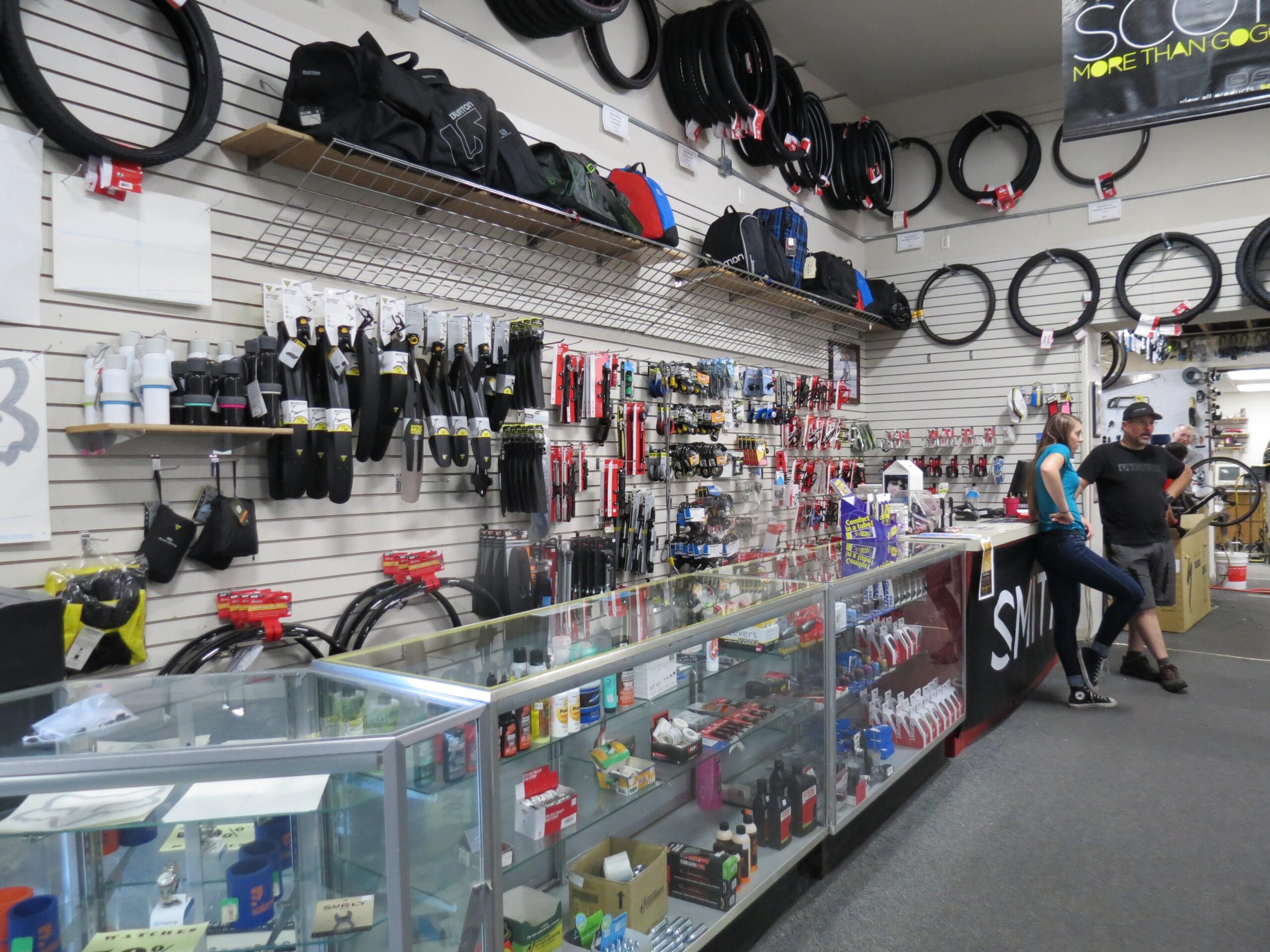 A bike shop interior featuring a display wall stocked with various cycling accessories, tools, and tires. The foreground includes a glass display case filled with bottles and bike care products. Two customers, a man and a woman, are casually engaging near the checkout area. The shop has a welcoming atmosphere with organized merchandise neatly arranged on shelves.