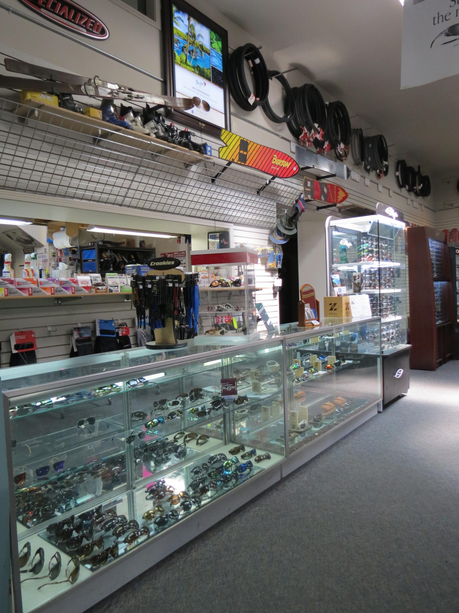 A display area in a retail store featuring a variety of sunglasses arranged in glass display cases. The background showcases shelves with ski equipment and other outdoor gear. A monitor displaying a tropical image hangs on the wall, while additional merchandise, including bike tires, is visible above the displays. The overall setting conveys an outdoor sports theme.