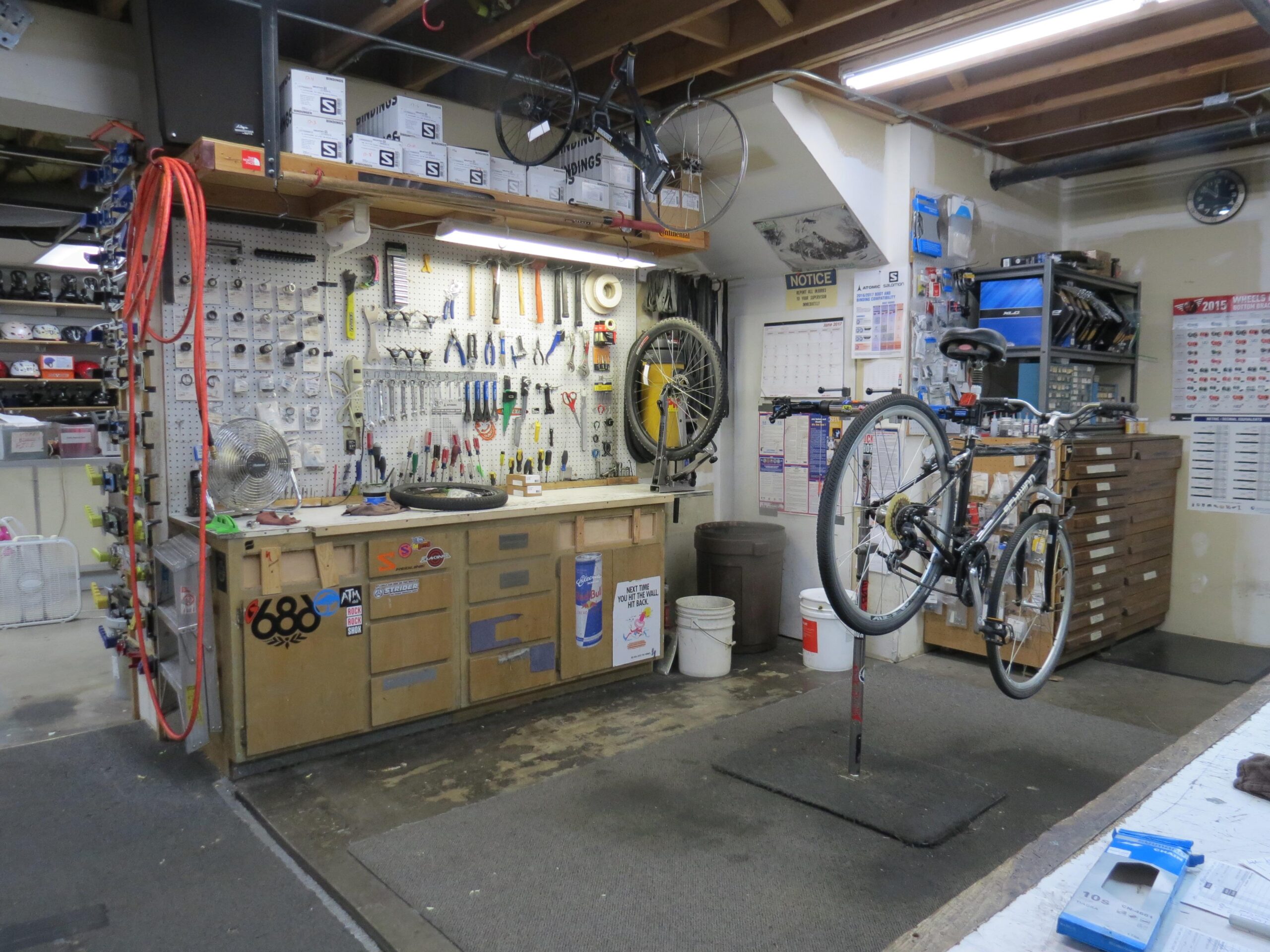 A bicycle is being serviced on a repair stand in a workshop filled with tools and equipment. The wall features a pegboard with various hand tools organized on hooks, while a workbench displays additional tools and a partially inflated bicycle tire. Shelving above contains storage boxes, and a calendar is visible on the wall. The space is well-lit and organized, reflecting a functional bike repair environment.