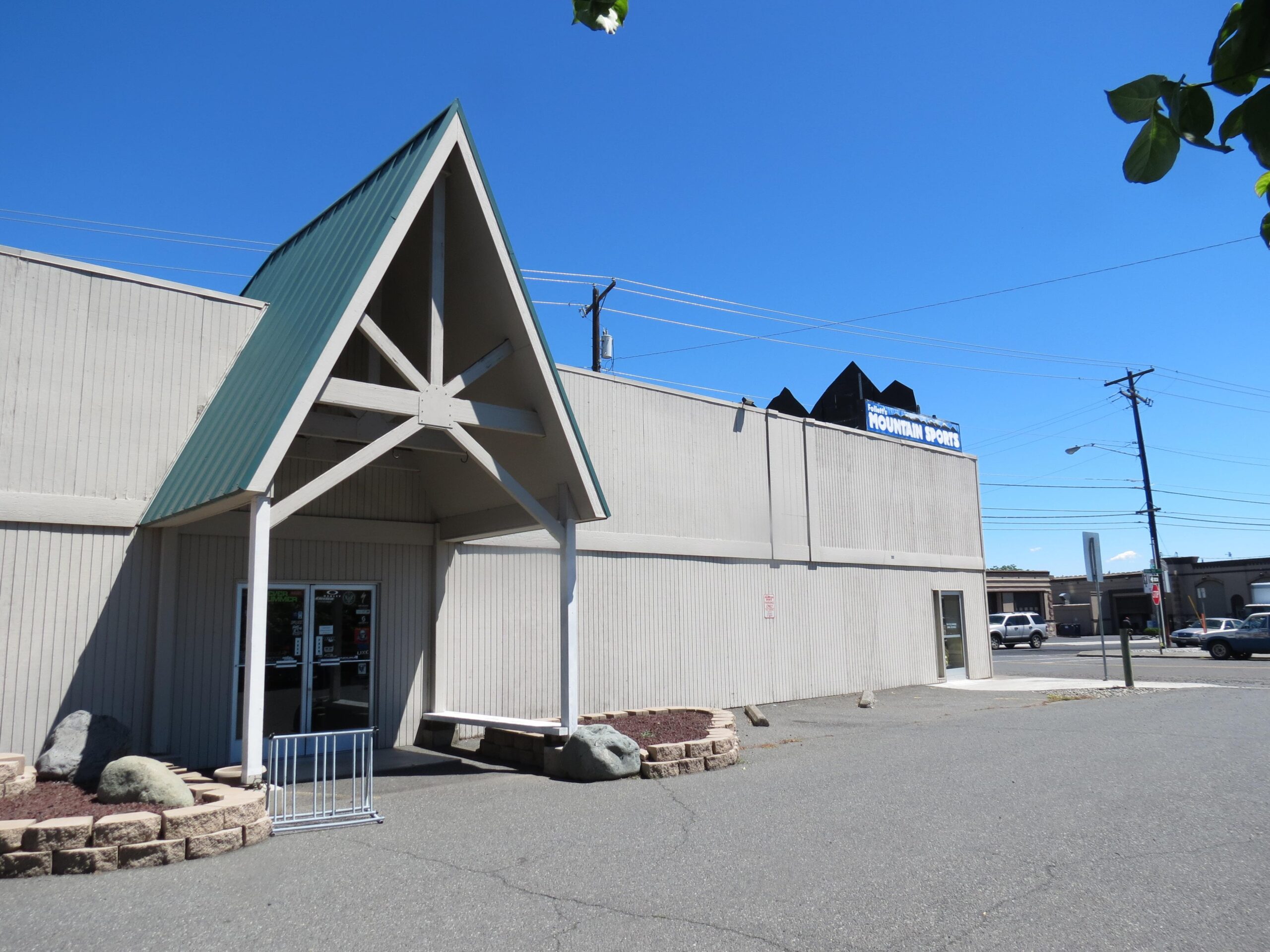 Exterior view of a building featuring a triangular roof structure and a sign for "Mountain Sports." The entrance is framed by light-colored siding, with a stone planter in front and a blue sky above.