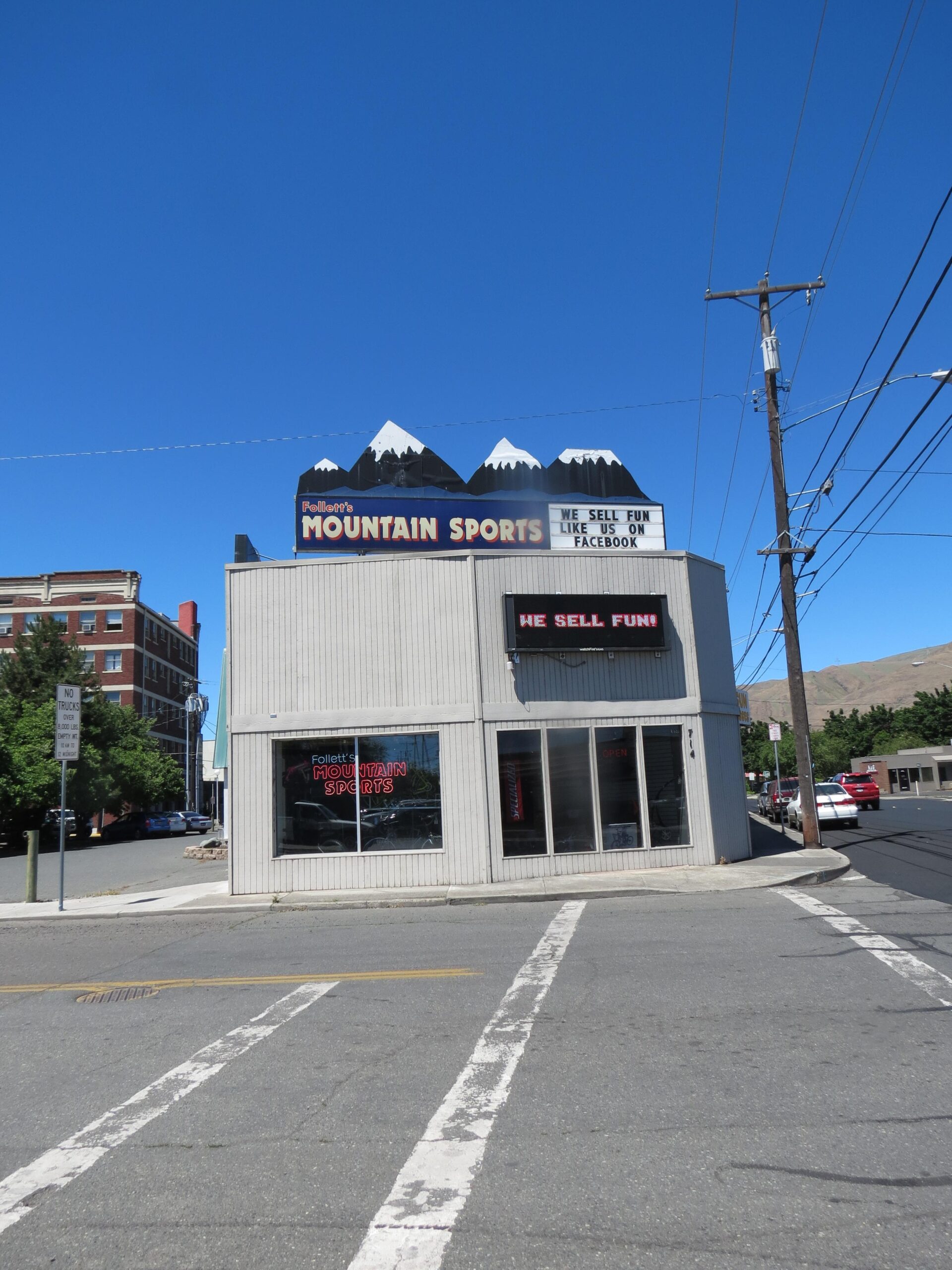 A corner building displaying a sign for "Follett's Mountain Sports," featuring stylized mountains with snow caps atop the roof. The storefront has large windows, with a neon "OPEN" sign visible. A message on the marquee reads, "WE SELL FUN, LIKE US ON FACEBOOK." The sky is clear and blue, and the street has white crosswalk lines.