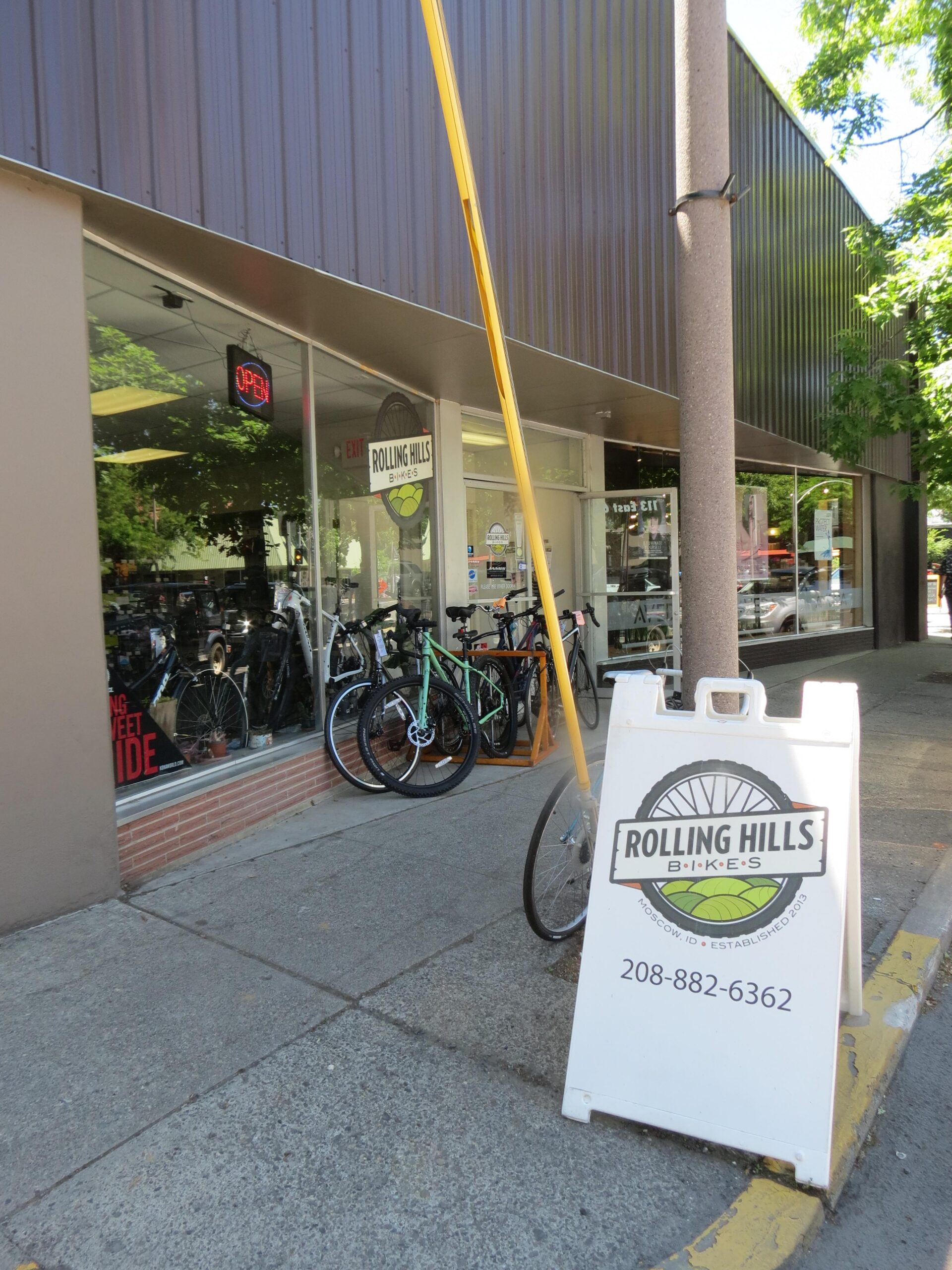 A storefront for Rolling Hills BIkes featuring a large window displaying various bicycles inside. An "Open" sign is illuminated, and a sidewalk sign provides the store's contact information. The exterior is well-lit and surrounded by greenery.