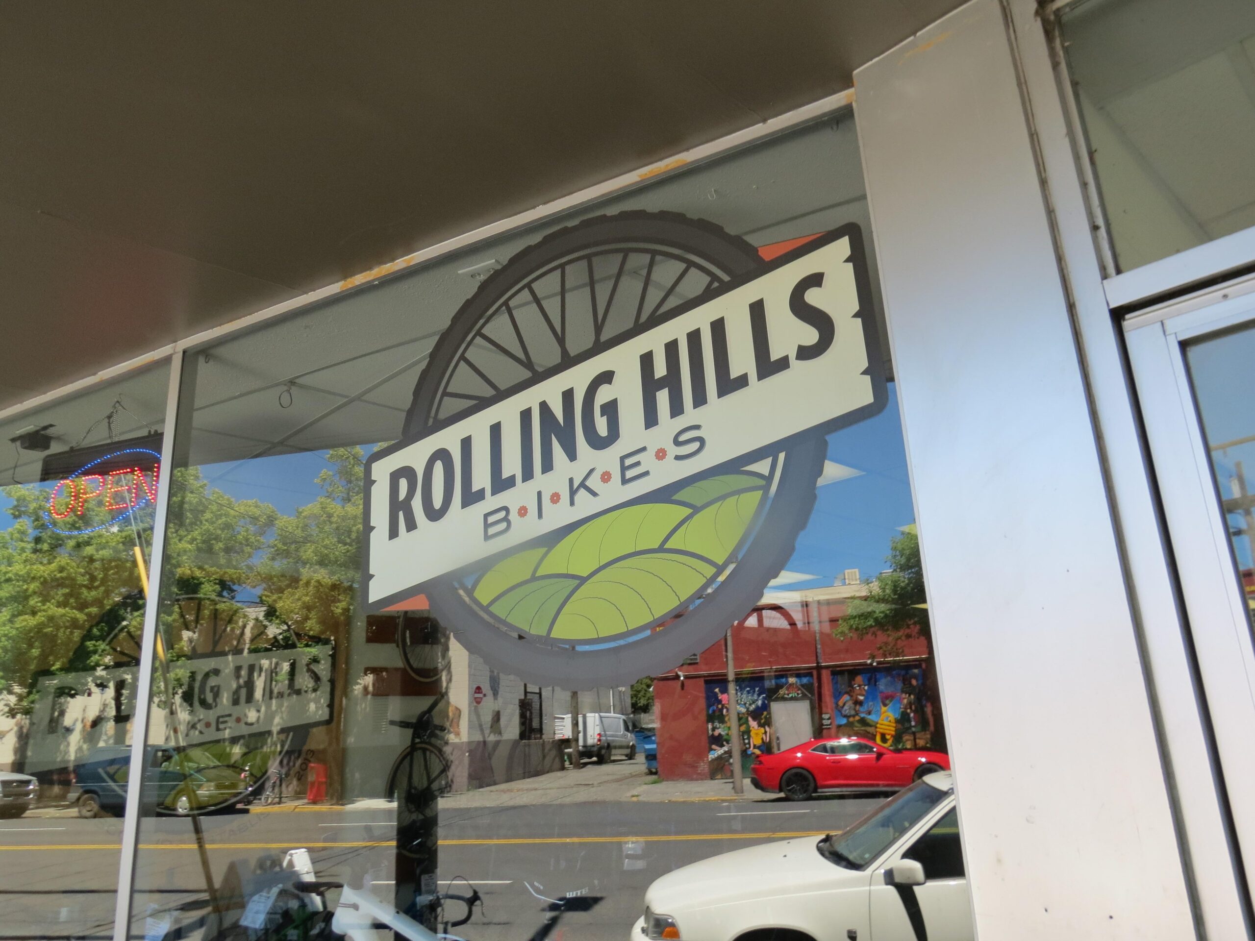 Alt text: "A storefront window featuring a sign for 'Rolling Hills Bikes,' with a visible bicycle wheel graphic. An illuminated 'OPEN' sign hangs above the window, reflecting a colorful mural and parked cars outside."