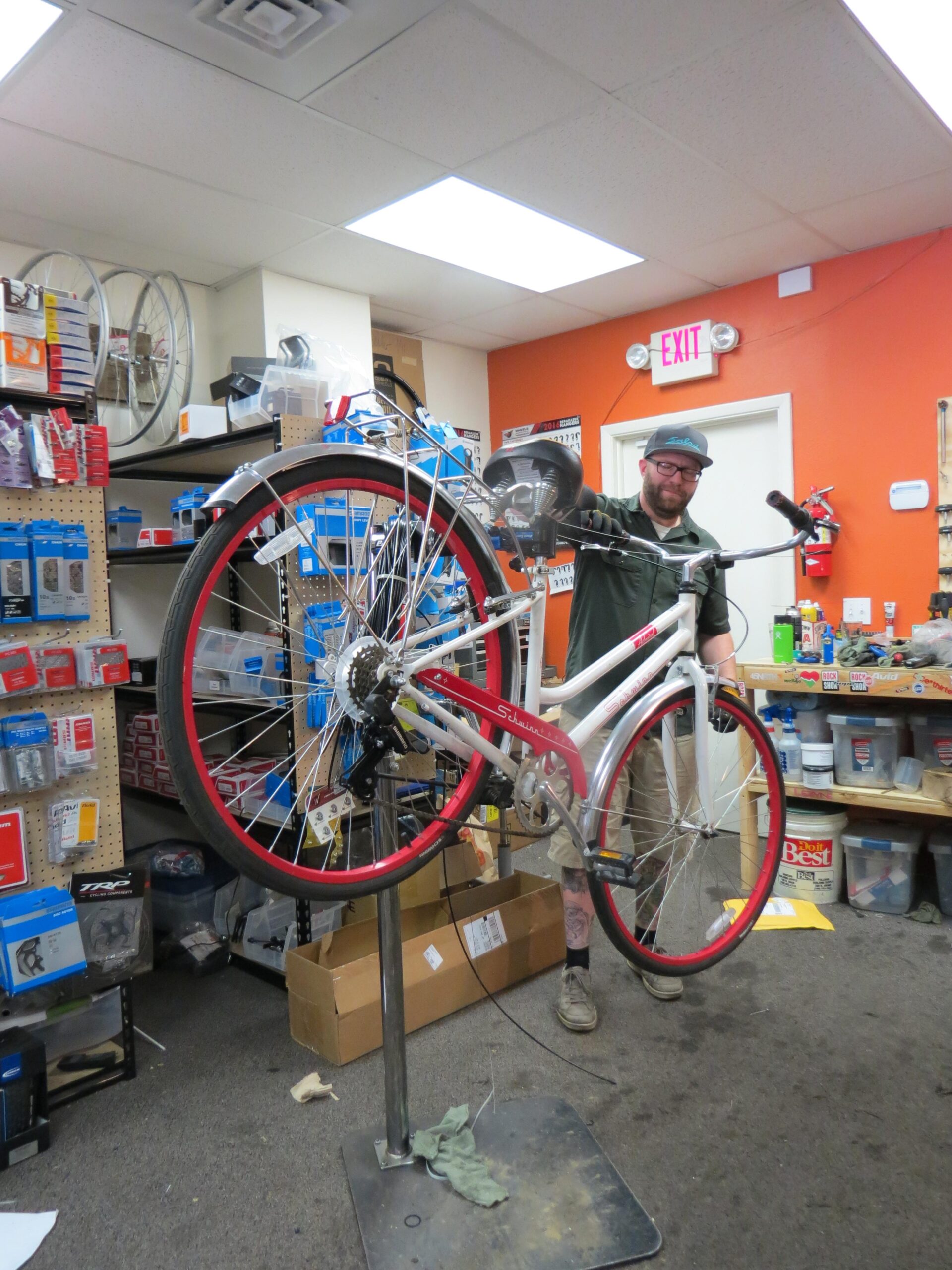 A mechanic is working on a red and white bicycle that is mounted on a repair stand inside a bike shop. The shop has shelves filled with various bike parts and accessories in the background, along with tools and supplies scattered around. Bright overhead lights illuminate the workspace.