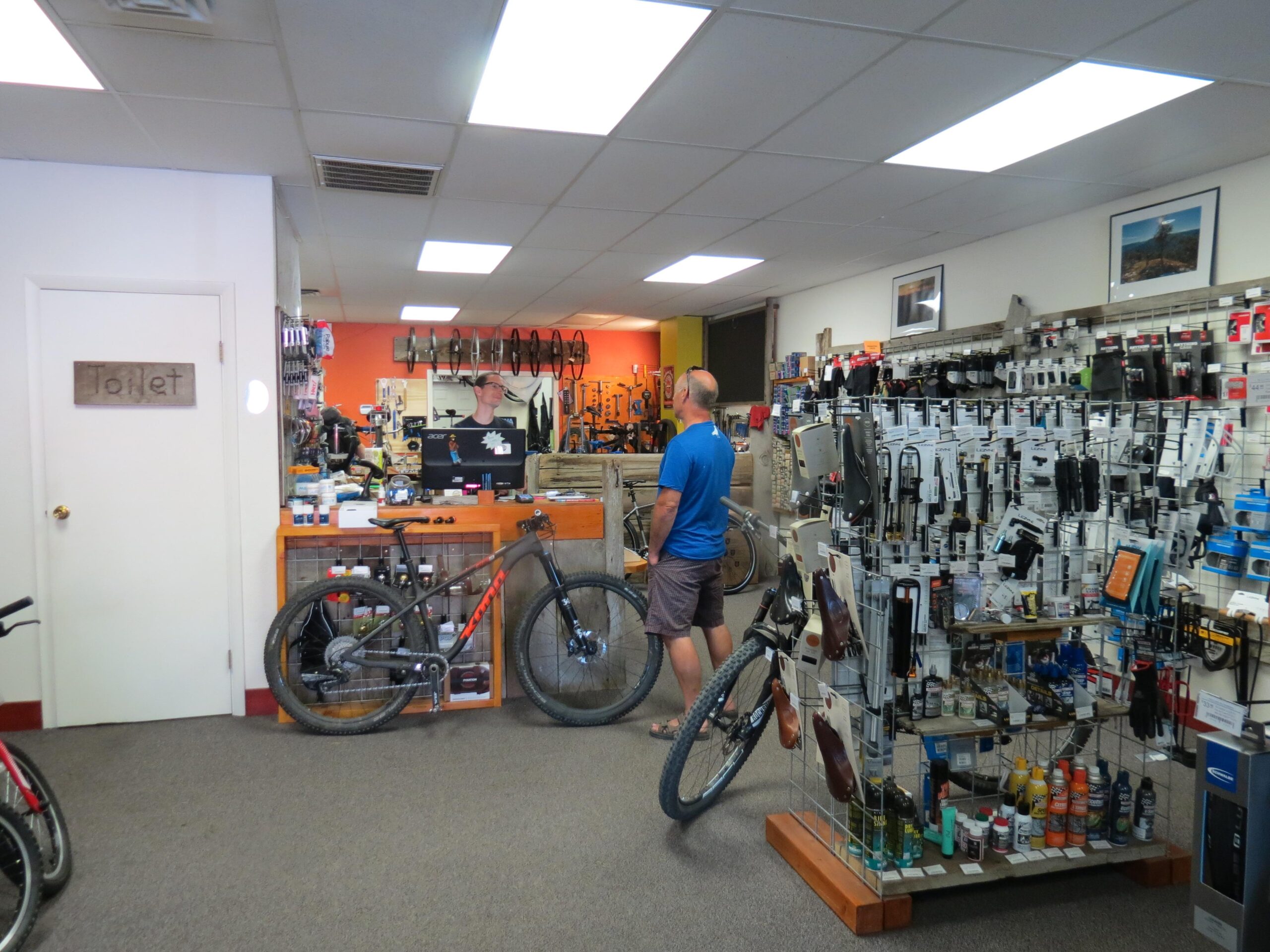 An interior view of a bike shop featuring various bicycles and cycling accessories. A man in a blue shirt stands near a display of bicycle parts and maintenance products, while a wooden sign indicates the location of the restroom. The shop has a vibrant orange wall and showcases different types of bikes.