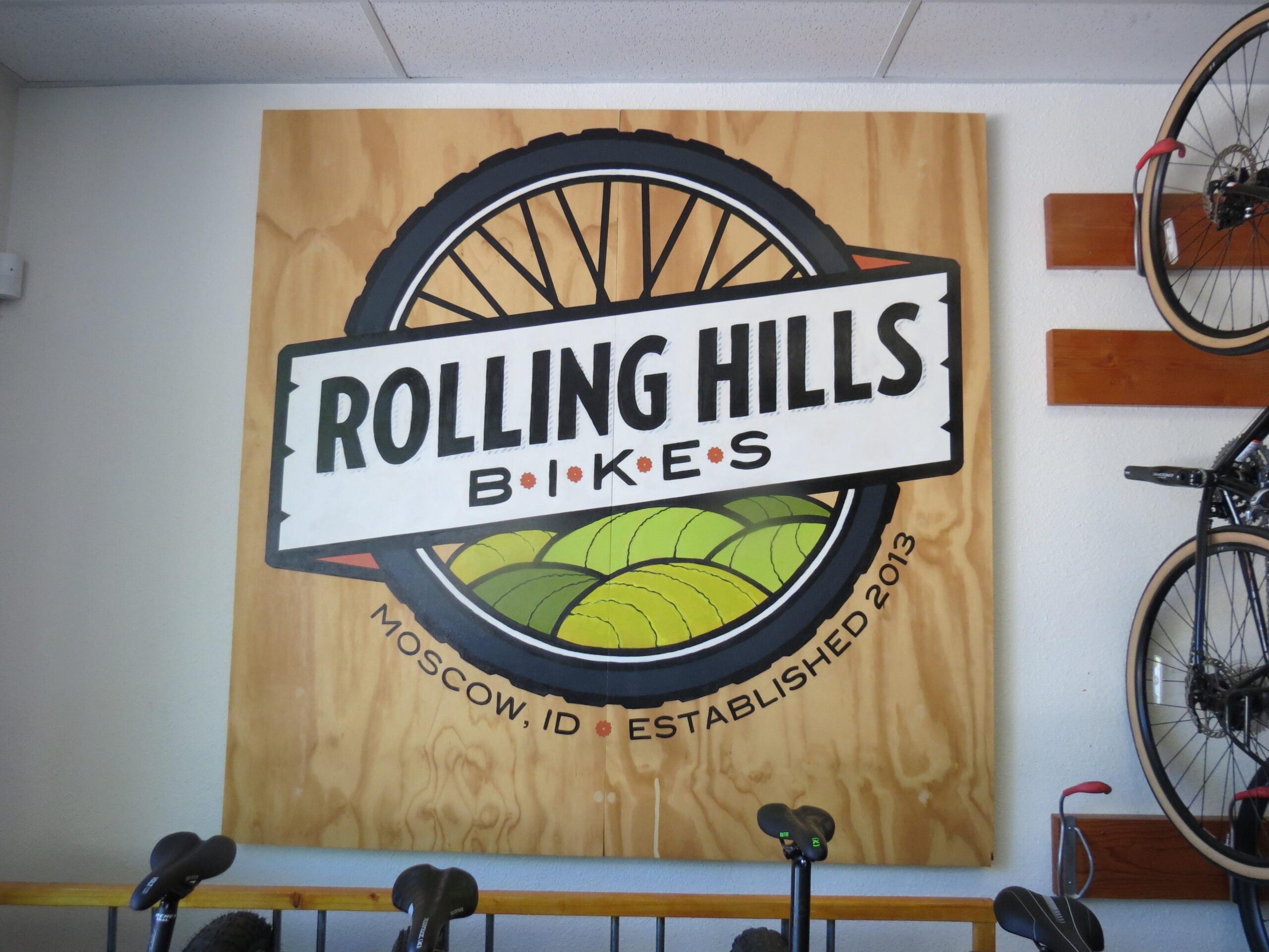 A wooden sign featuring the logo for "Rolling Hills Bikes" with a bicycle wheel design and green hills, located in Moscow, Idaho. The sign indicates the establishment year as 2013. In the background, bicycle parts and bikes are displayed on the wall.
