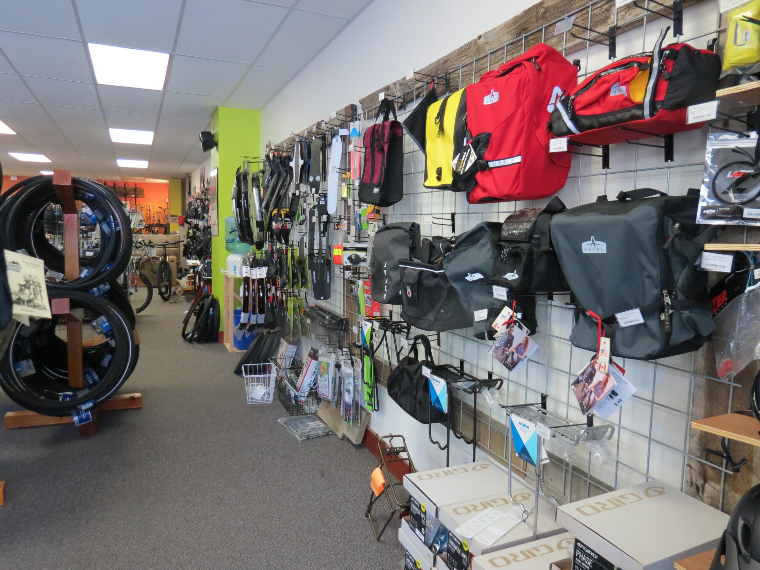 Image of a bicycle shop interior showcasing various bike accessories and equipment. The walls are lined with bike bags and gear, while the center features bicycle tires displayed on wooden stands. The space is well-lit and organized, with a variety of products available for cycling enthusiasts.