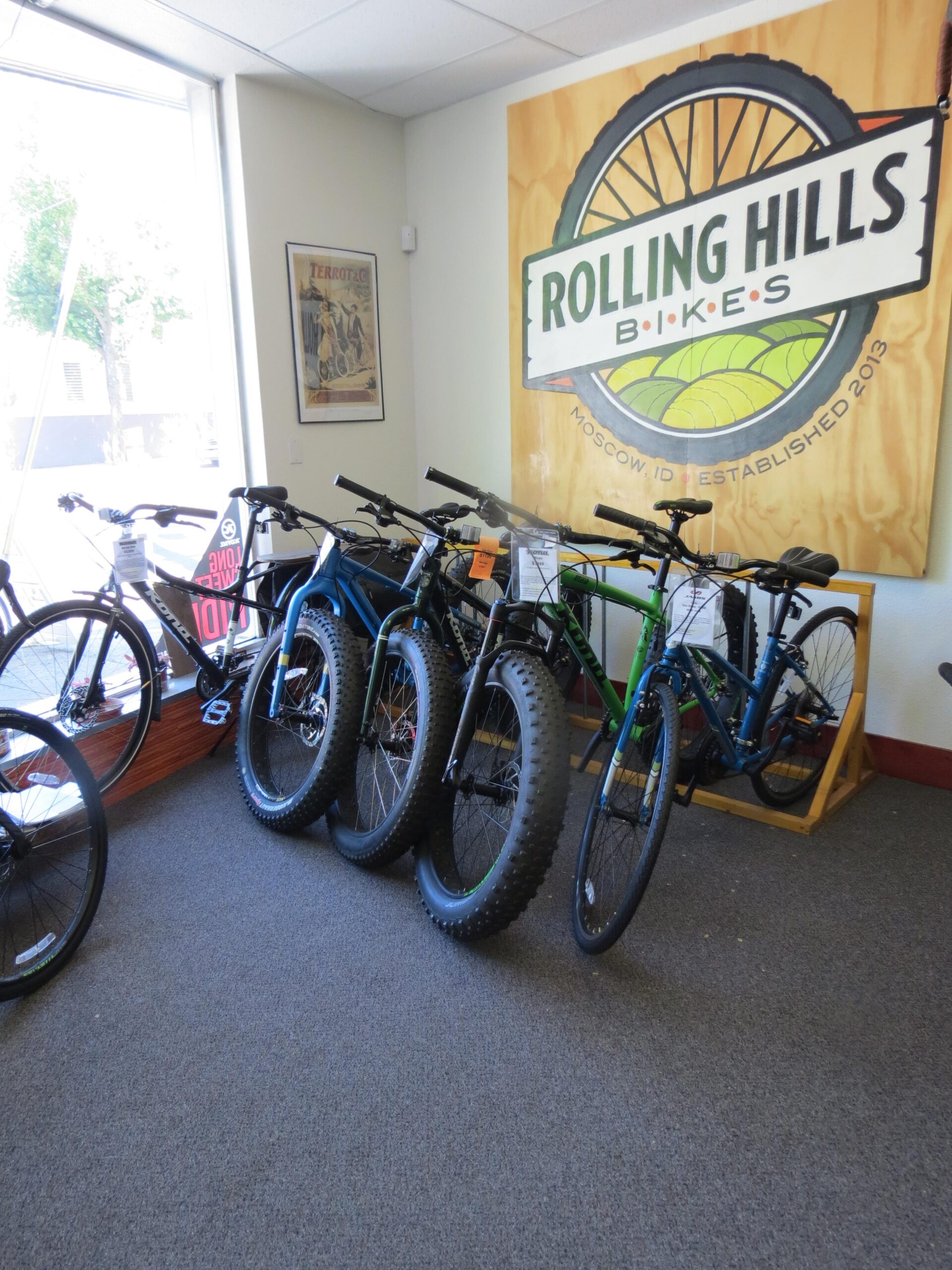Interior view of a bike shop featuring several bicycles against a wall with a large sign that reads "Rolling Hills Bikes." The shop includes a mix of regular bicycles and fat bikes, with various price tags attached. There is a window showing outdoor greenery, and a vintage cycling poster can be seen on the wall. The floor is carpeted, contributing to the welcoming atmosphere.