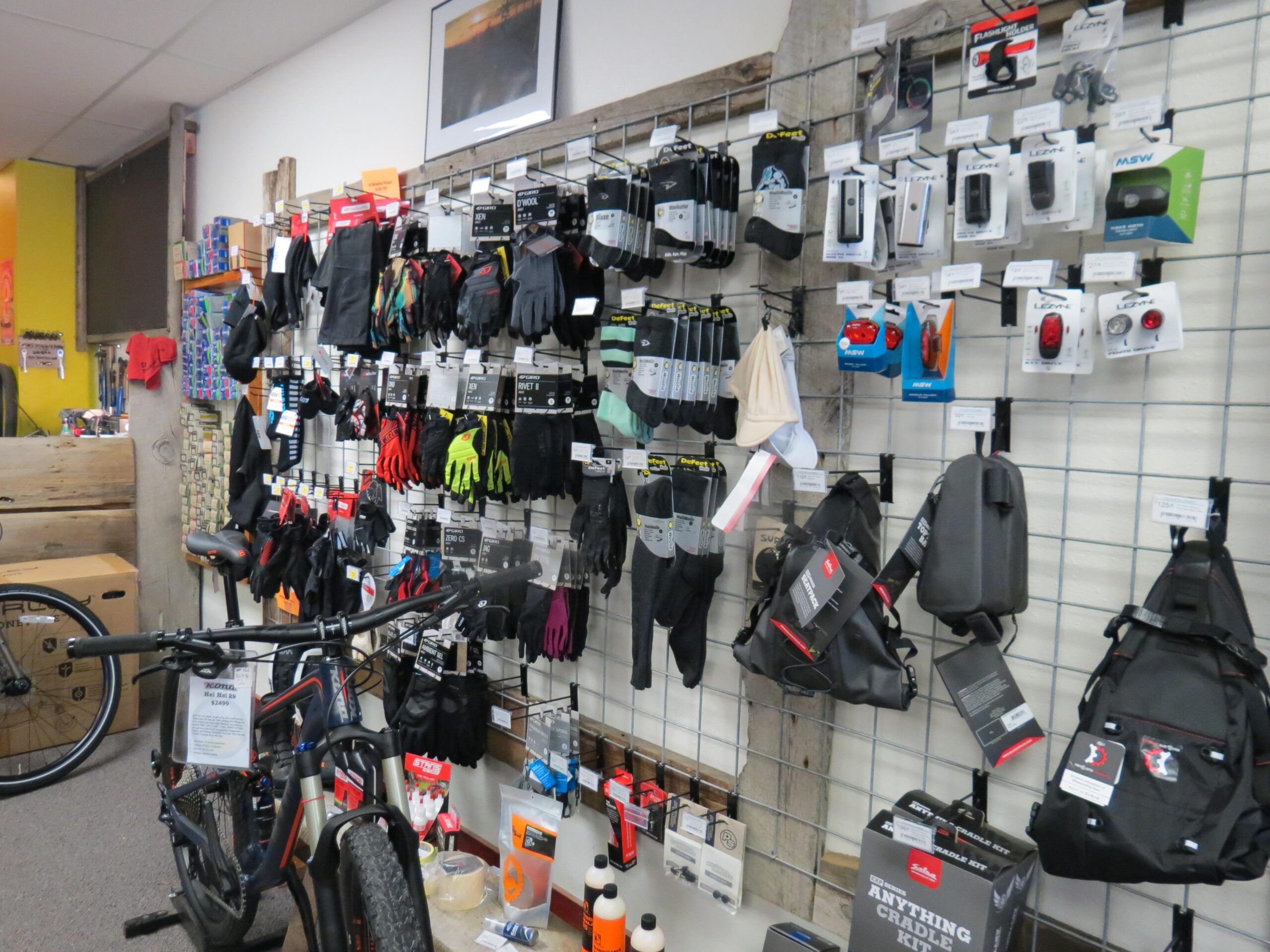 A retail display featuring a variety of cycling accessories, including gloves, bike lights, and bags. The items are organized on a grid wall, with labels and pricing visible. In the foreground, a mountain bike is partially visible, showcasing its handlebars and front wheel. The background includes a colorful arrangement of additional merchandise and a framed photo on the wall.