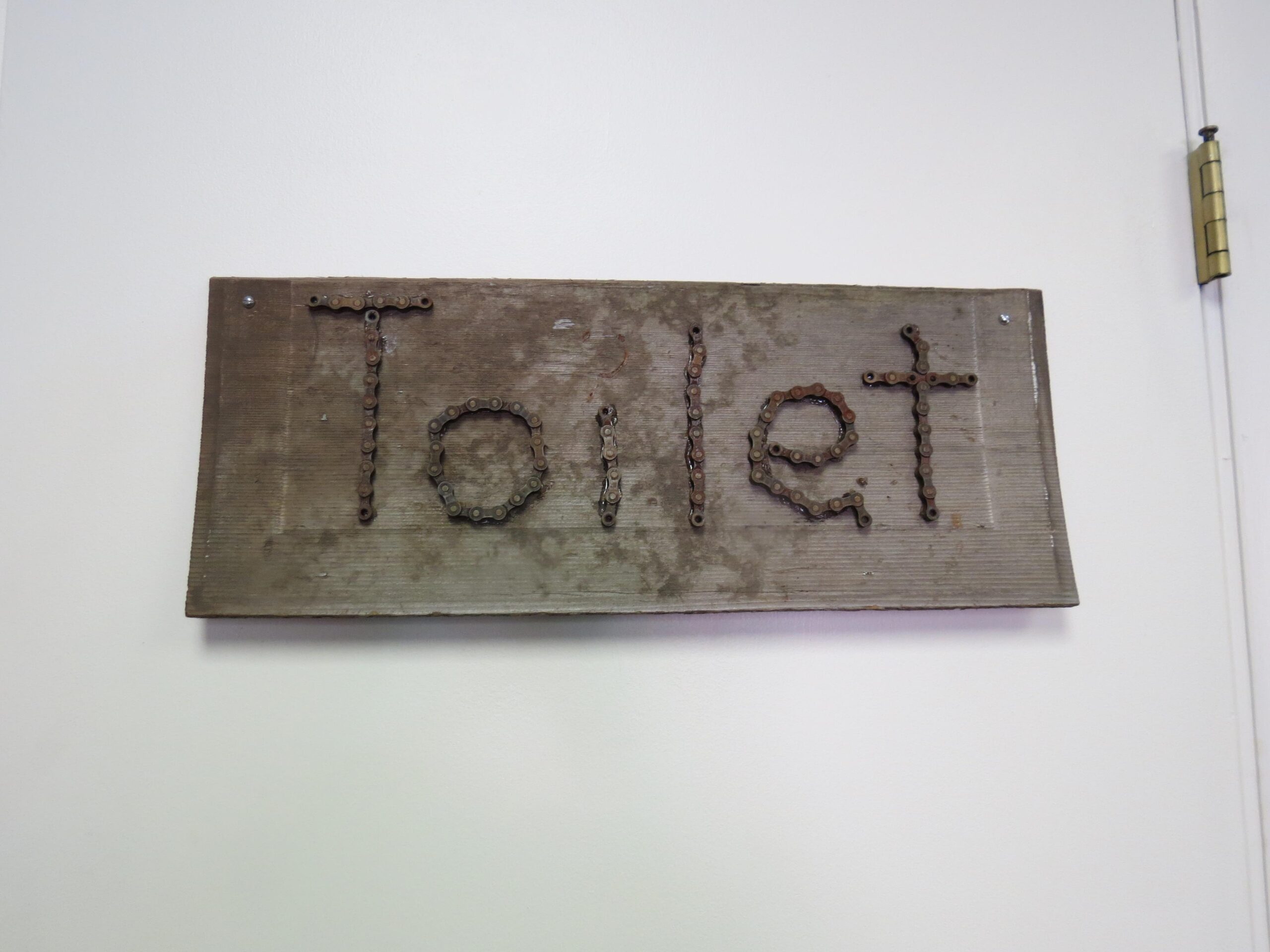 A wooden sign with the word "Toilet" crafted from what appears to be metal links, mounted on a wall. The surface of the wood is slightly weathered, and the sign is attached with screws.