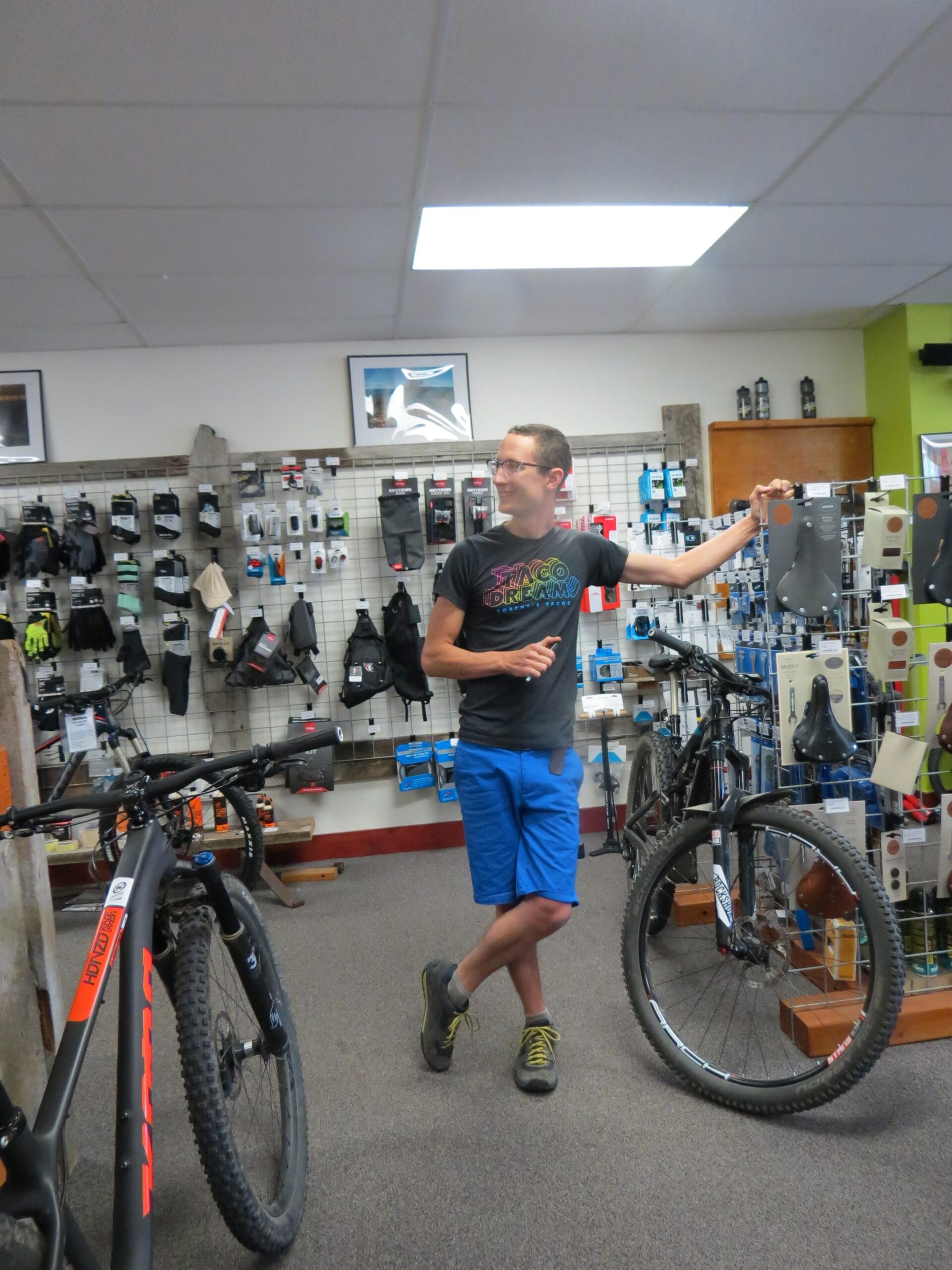 A person stands in a bicycle shop, leaning against a display with one hand on a bicycle component. They are wearing a colorful t-shirt and blue shorts, surrounded by mounted bicycles and various cycling accessories. The shop has a well-organized layout with products visible on the walls.