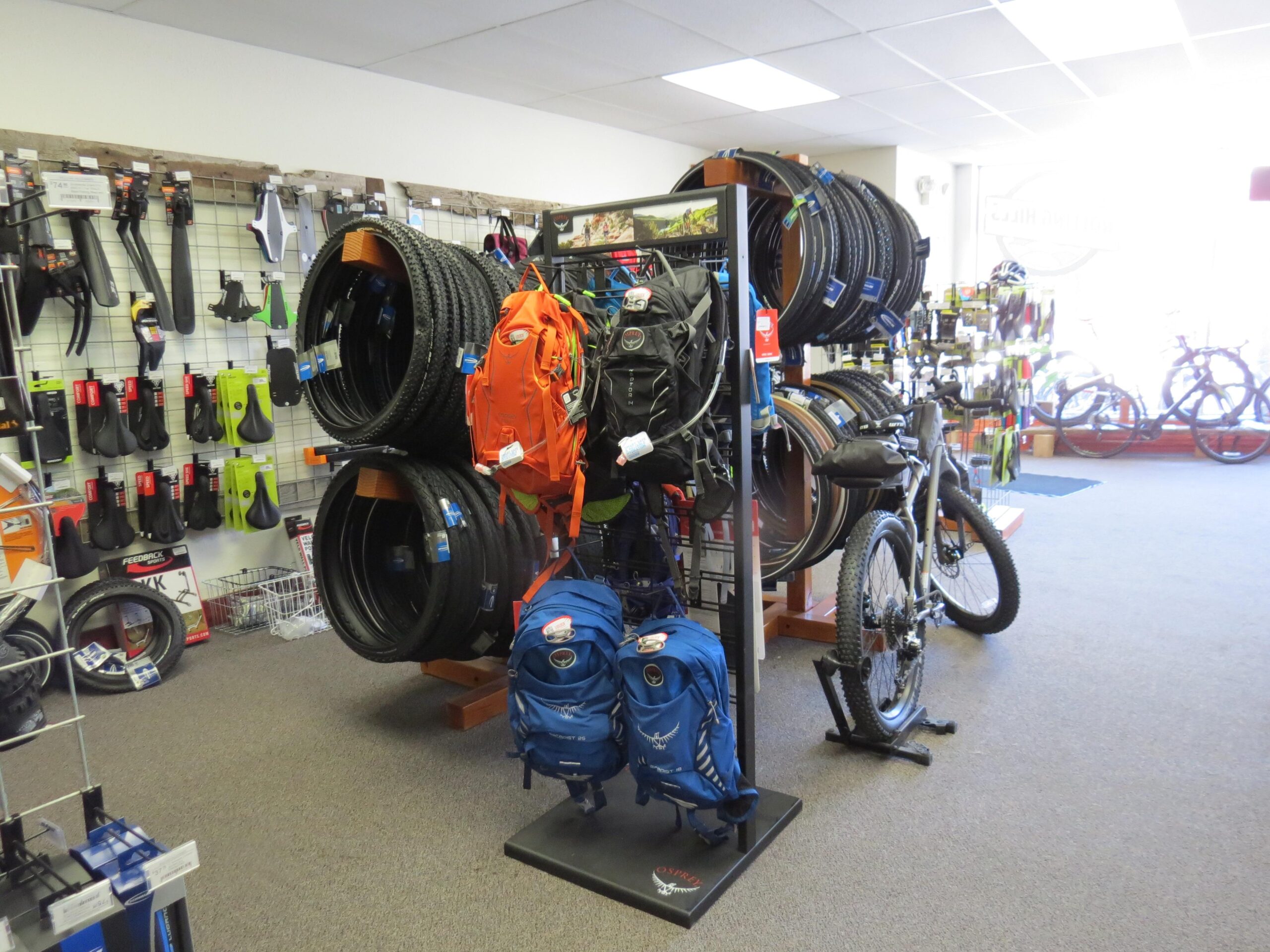 A brightly lit bicycle shop interior featuring a variety of biking accessories. On display are bicycle tires, backpacks, and other gear on shelves and racks. A mountain bike is positioned prominently in the foreground, while more bicycles can be seen in the background. The shop has a clean and organized appearance, with various colored items contributing to a vibrant atmosphere.