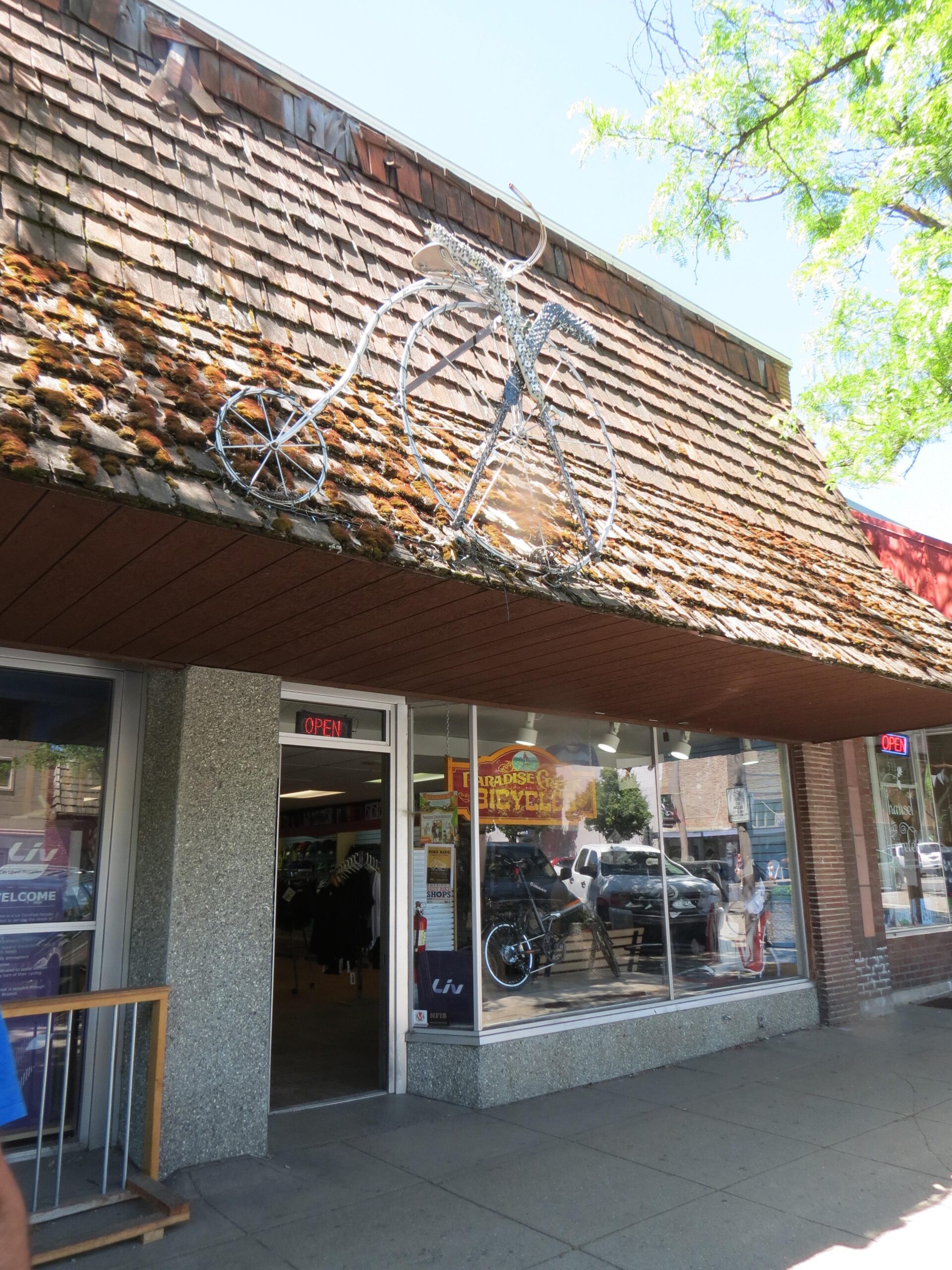 A bicycle-themed display featuring a metal sculpture of a bicycle on the roof of a storefront. The store, which appears to be a bicycle shop, has glass windows with various bicycles and merchandise visible inside. The entrance is marked with an "OPEN" sign, and the building has a charming, rustic wooden exterior with a shingled roof.