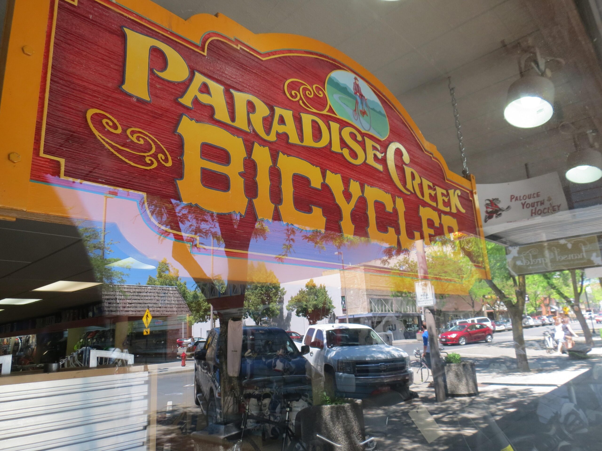A colorful sign reading "Paradise Creek Bicycles" hanging in a shop window, reflecting the vibrant street scene outside, which includes cars, trees, and pedestrians.