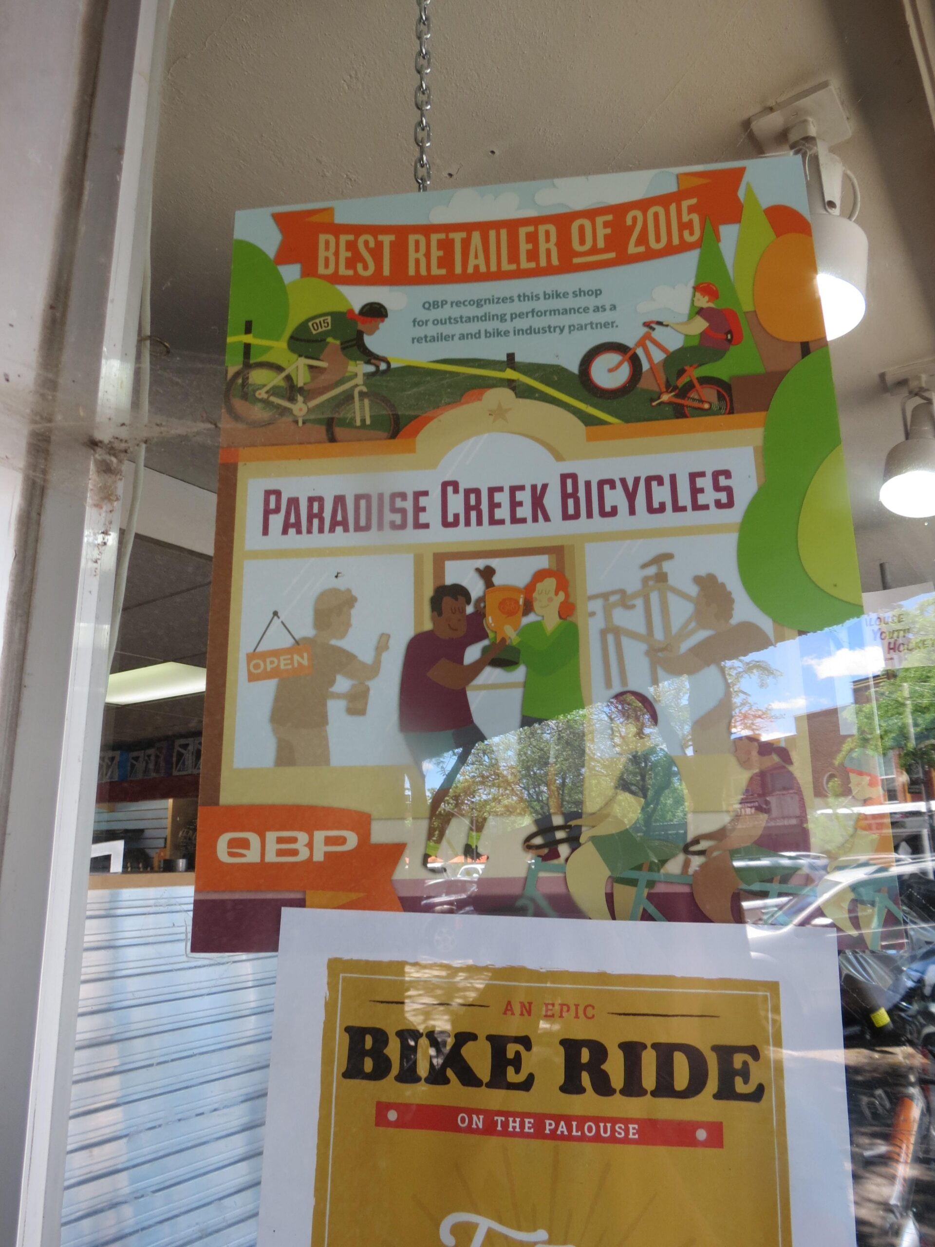 A colorful poster displayed in a bike shop window, promoting Paradise Creek Bicycles as the "Best Retailer of 2015" according to QBP. The design features illustrations of cyclists and cheerful figures enjoying biking-related activities, along with an "OPEN" sign. Below the main poster, there is an additional sign advertising an "Epic Bike Ride on the Palouse."