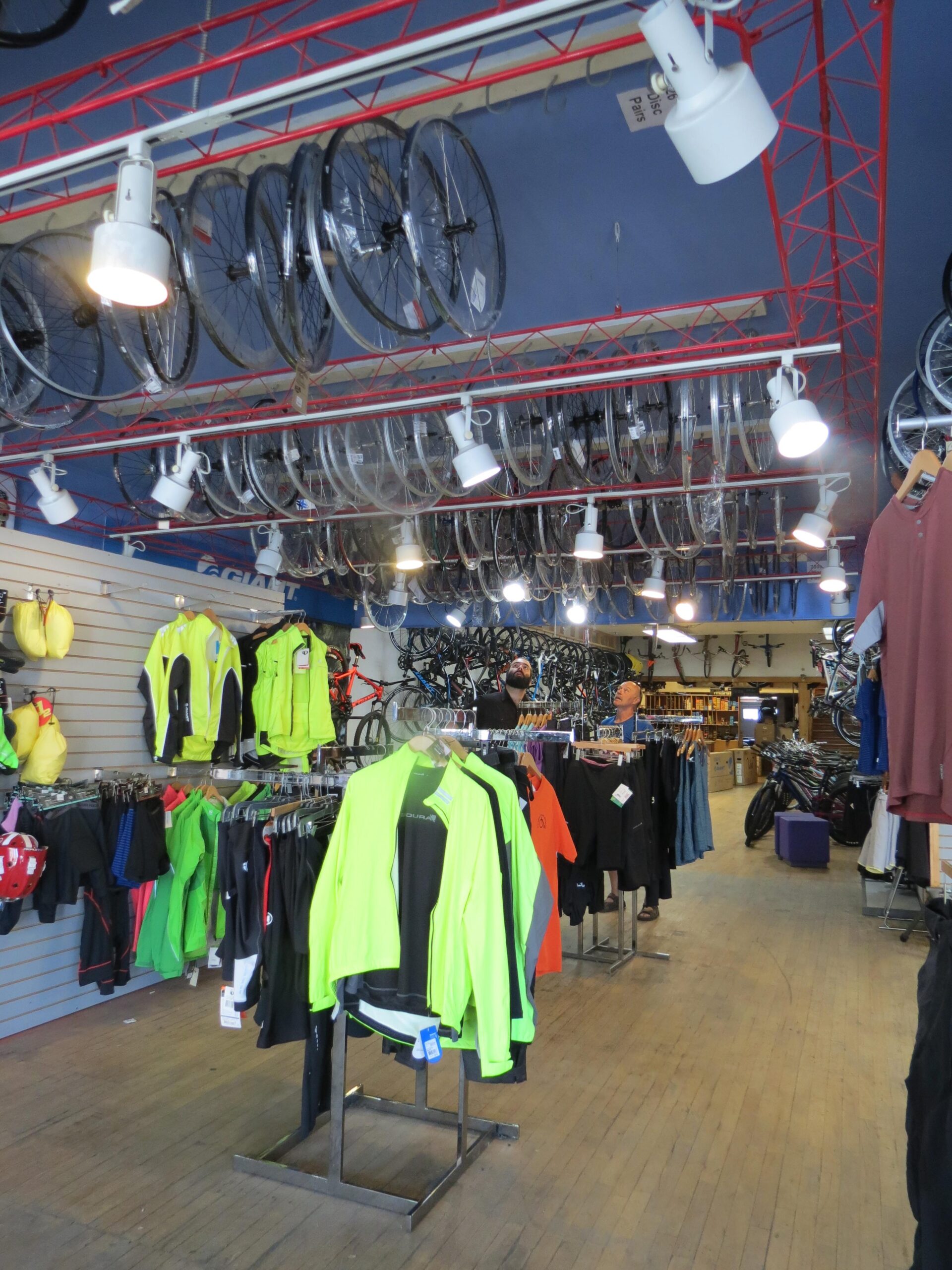 A spacious bike shop with colorful cycling apparel displayed on racks. Brightly colored jackets, jerseys, and other clothing items hang on the left side, while bicycle wheels are suspended from the ceiling. In the background, various bicycles are visible, and two customers are browsing. The shop has a warm ambiance with wooden flooring and well-lit fixtures.
