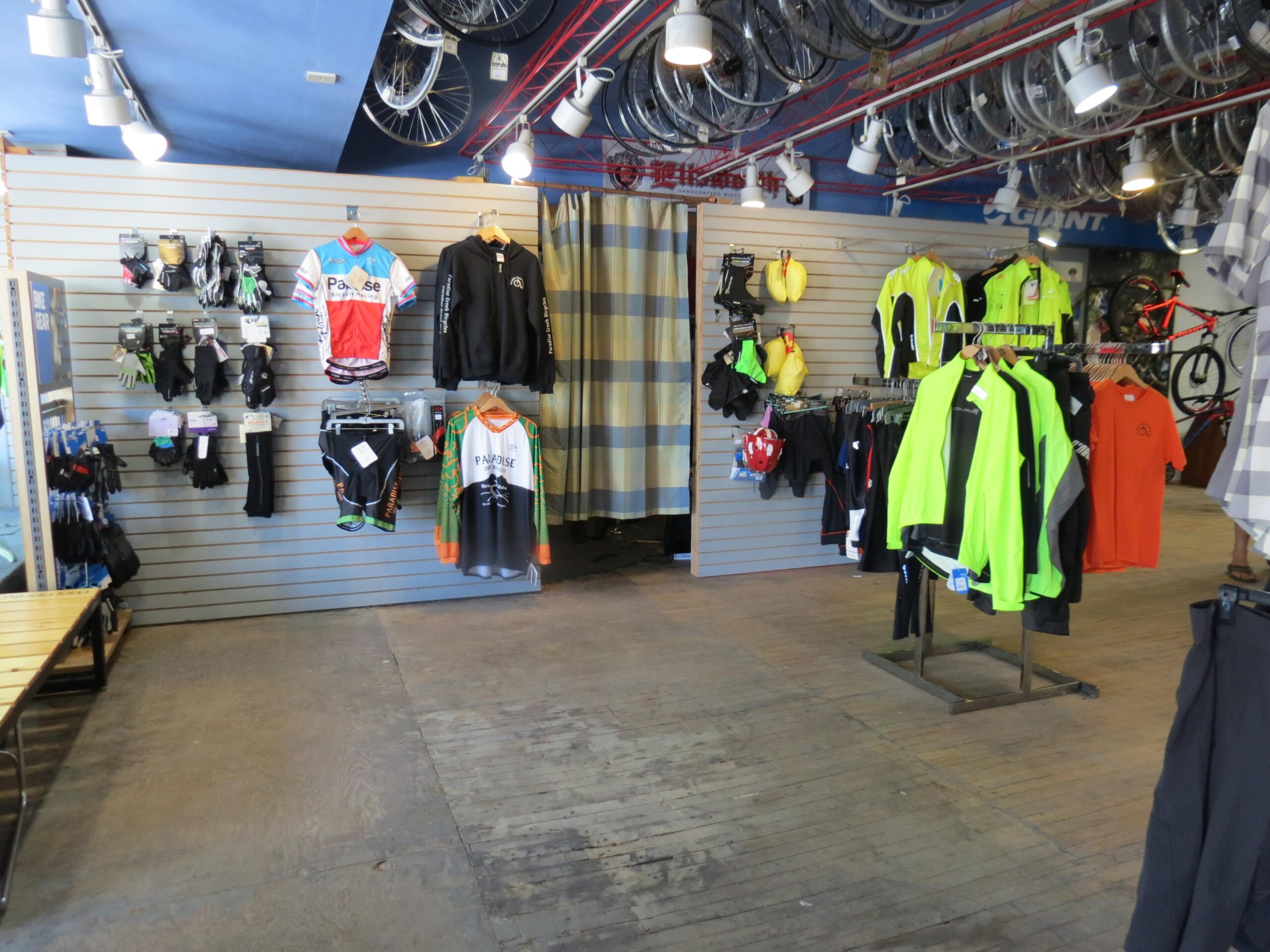 Image of an indoor cycling shop displaying various cycling apparel and accessories. On the left side, there are gloves, shorts, and jerseys hanging on the wall. In the center, a stand holds several brightly colored cycling jackets, including neon yellow options. The flooring is wooden, and bicycles can be seen hanging from the ceiling above. The overall layout suggests a well-organized retail space focused on cycling gear.