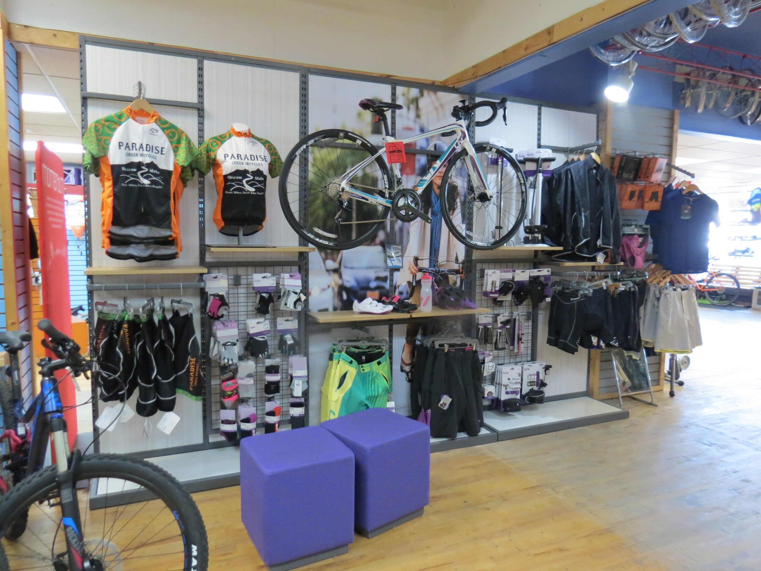 Shop interior featuring a display of cycling apparel and accessories. The wall showcases various cycling jerseys with "Paradise" branding, alongside a range of biking gear, including shorts and gloves. A bicycle is showcased above the merchandise, with additional cycling clothing hanging nearby. Two purple stools are positioned in front of the display on a wooden floor.