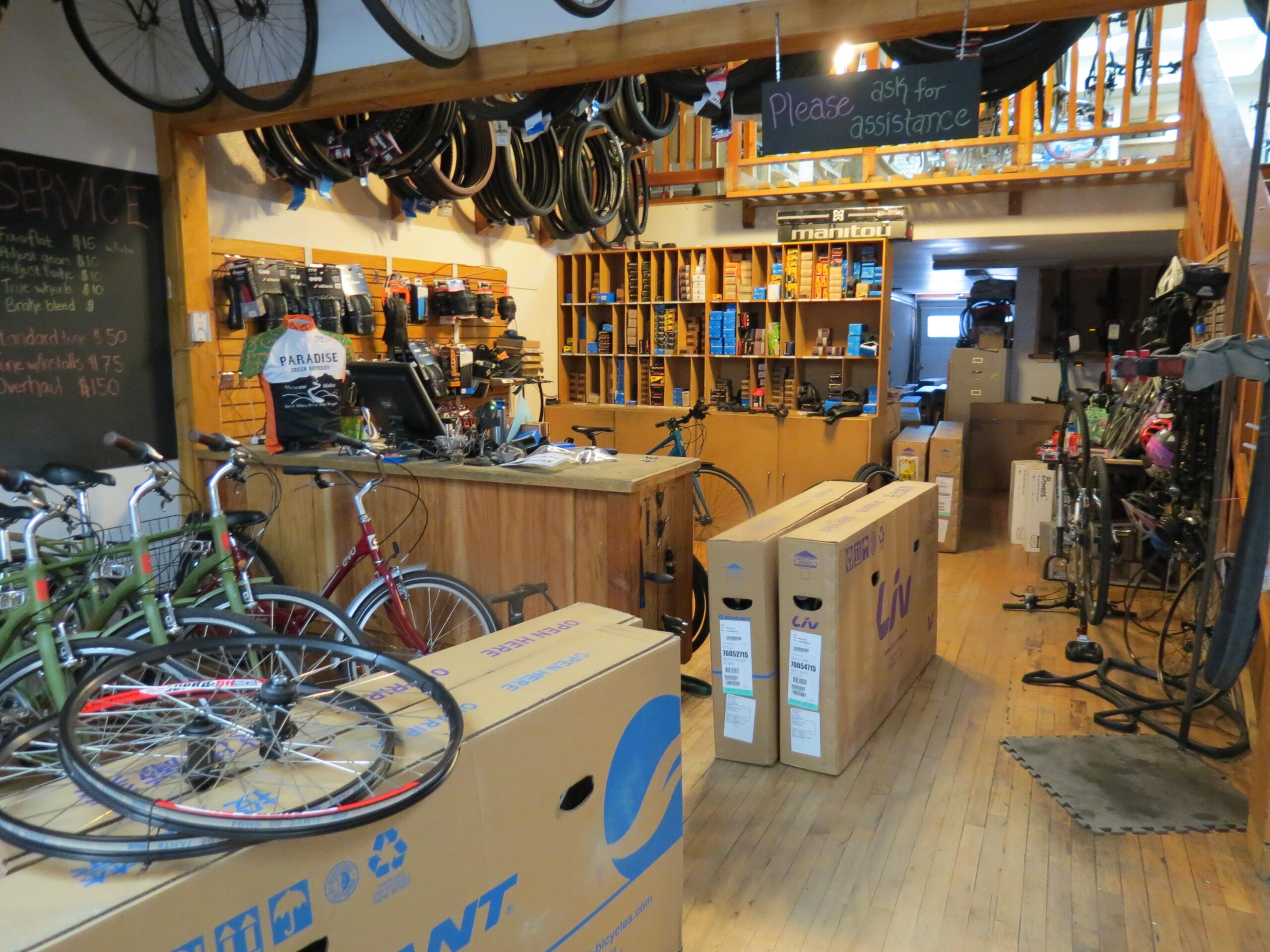 A cozy bicycle shop interior featuring various bikes, a wooden counter with a cash register, and shelves stocked with bike accessories. In the foreground, several bikes are leaning against large cardboard boxes. The walls display bike tires and a chalkboard listing services and prices. A sign overhead requests customers to ask for assistance.