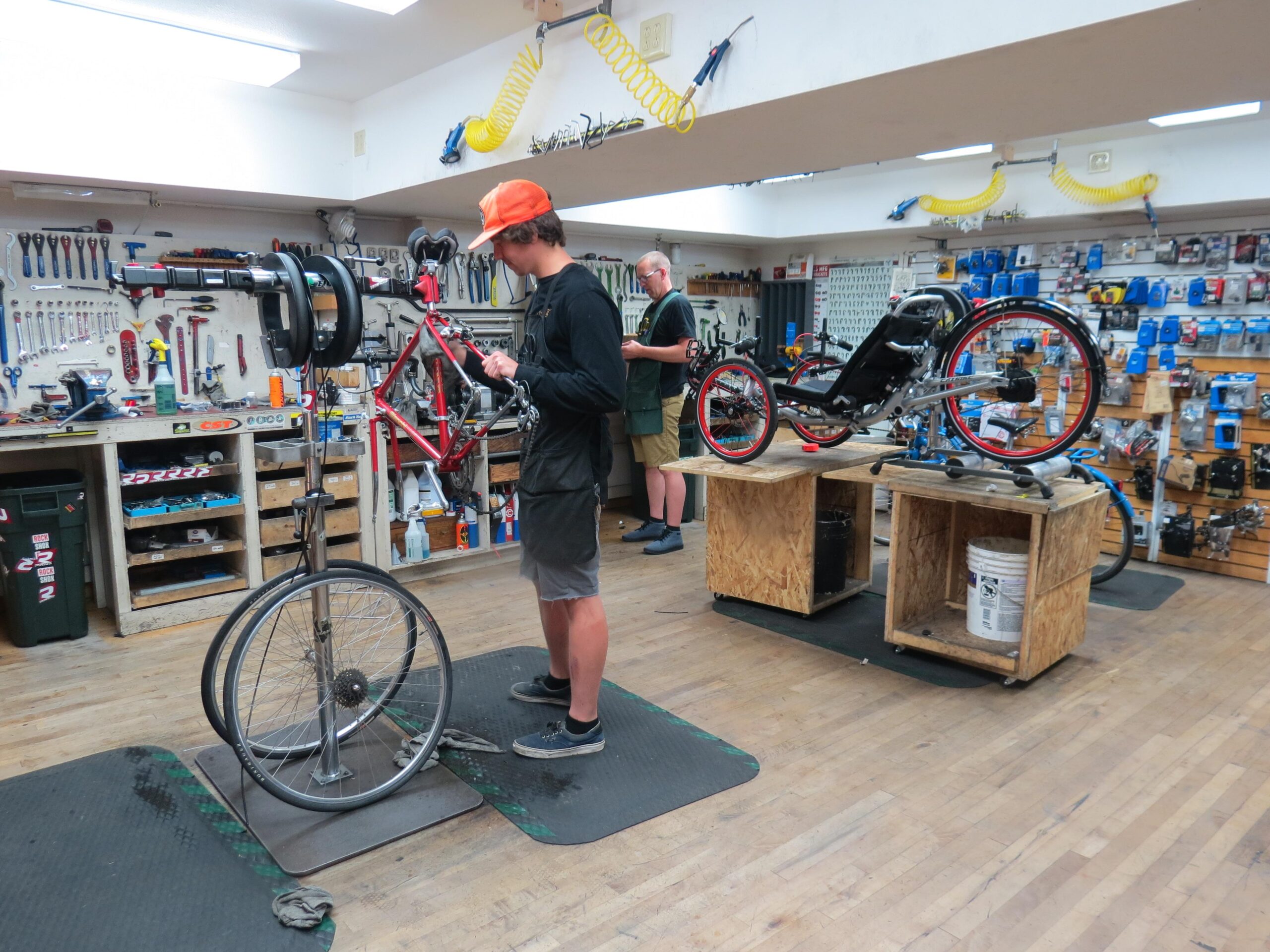 A bike repair shop interior featuring two individuals working on bicycles. One person, wearing a black long-sleeve shirt and an orange cap, is repairing a red bicycle on a stand, while another individual in the background, dressed in a green apron, is working on a recumbent bike displayed on a wooden table. The walls are lined with various bike tools and accessories. The floor is wooden, and there are toolboxes and materials scattered around.