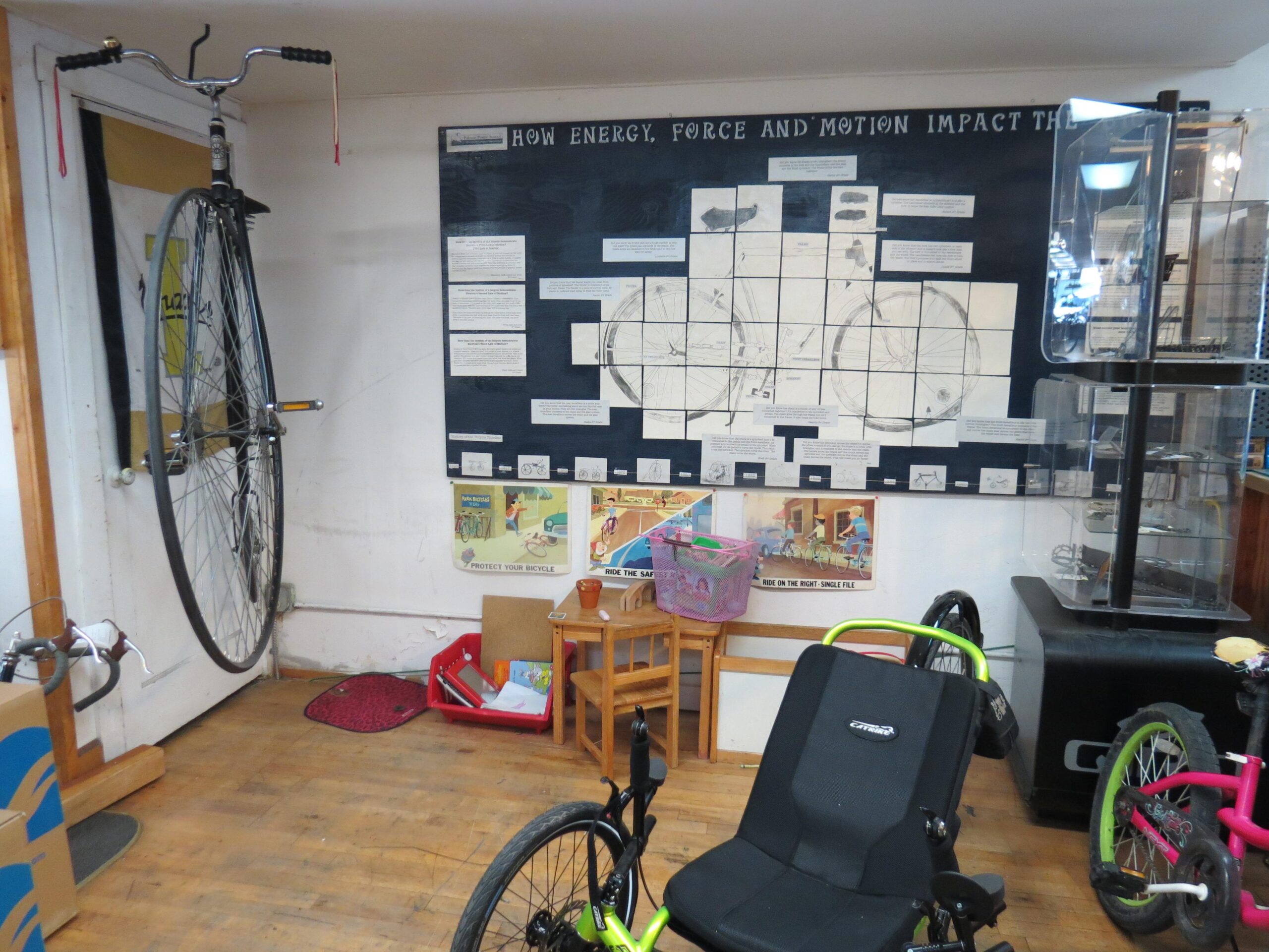 An indoor space featuring a vintage high-wheel bicycle mounted on the wall, accompanied by an educational display titled "How Energy, Force and Motion Impact the Bicycle." The area includes a small table and chairs, a colorful basket, and various bicycles, including a recumbent bike. The wooden floor adds warmth to the environment.
