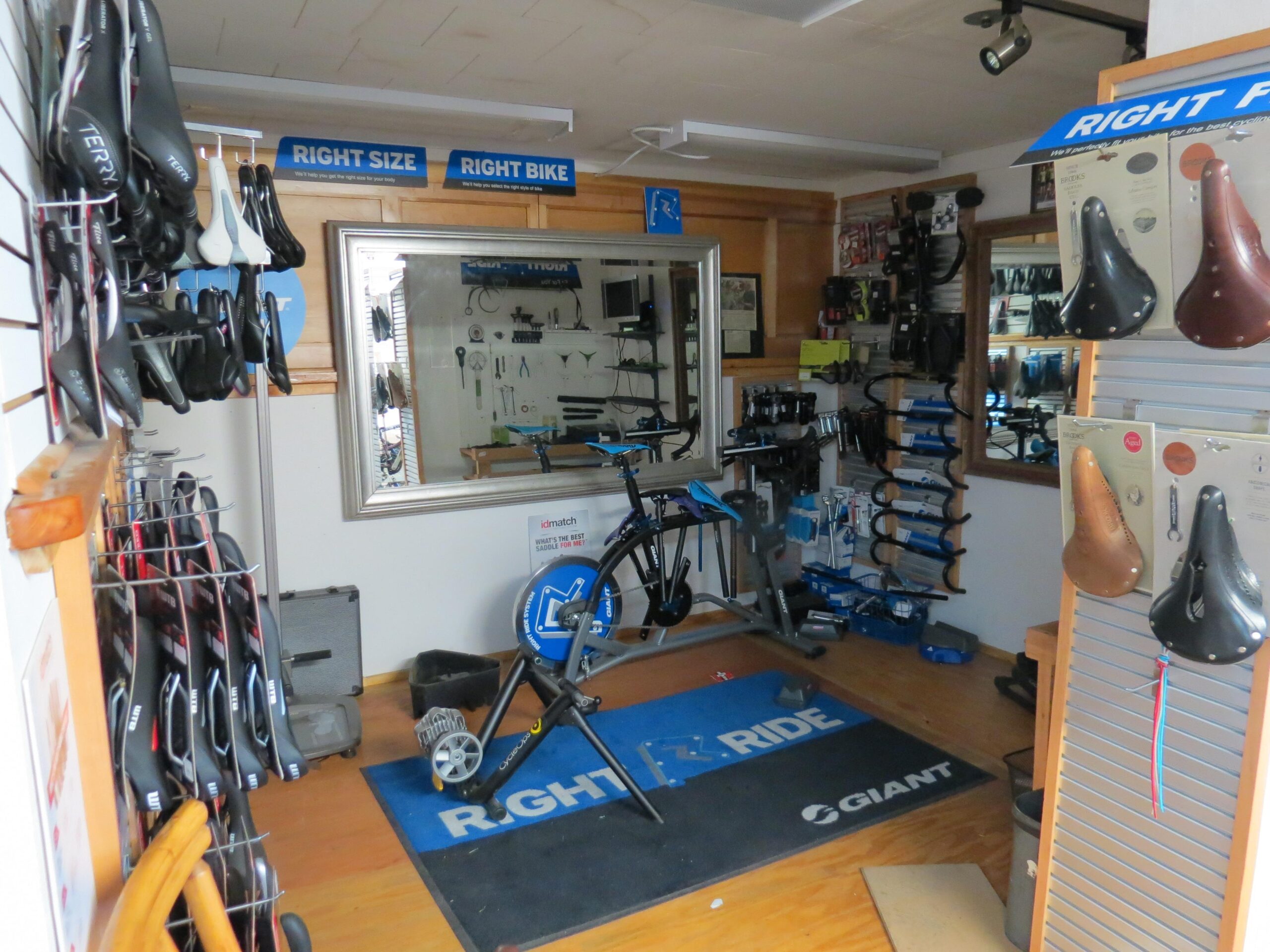 A bicycle shop interior featuring various bike saddles displayed on the walls and racks. In the center, a bike fitting station is visible with tools and equipment organized on shelves. A mirror is positioned on the back wall, and signs promoting "Right Size" and "Right Bike" are hanging above, emphasizing bike fitting services. The flooring has a branded mat with "RIGHT RIDE" and "GIANT" logos.