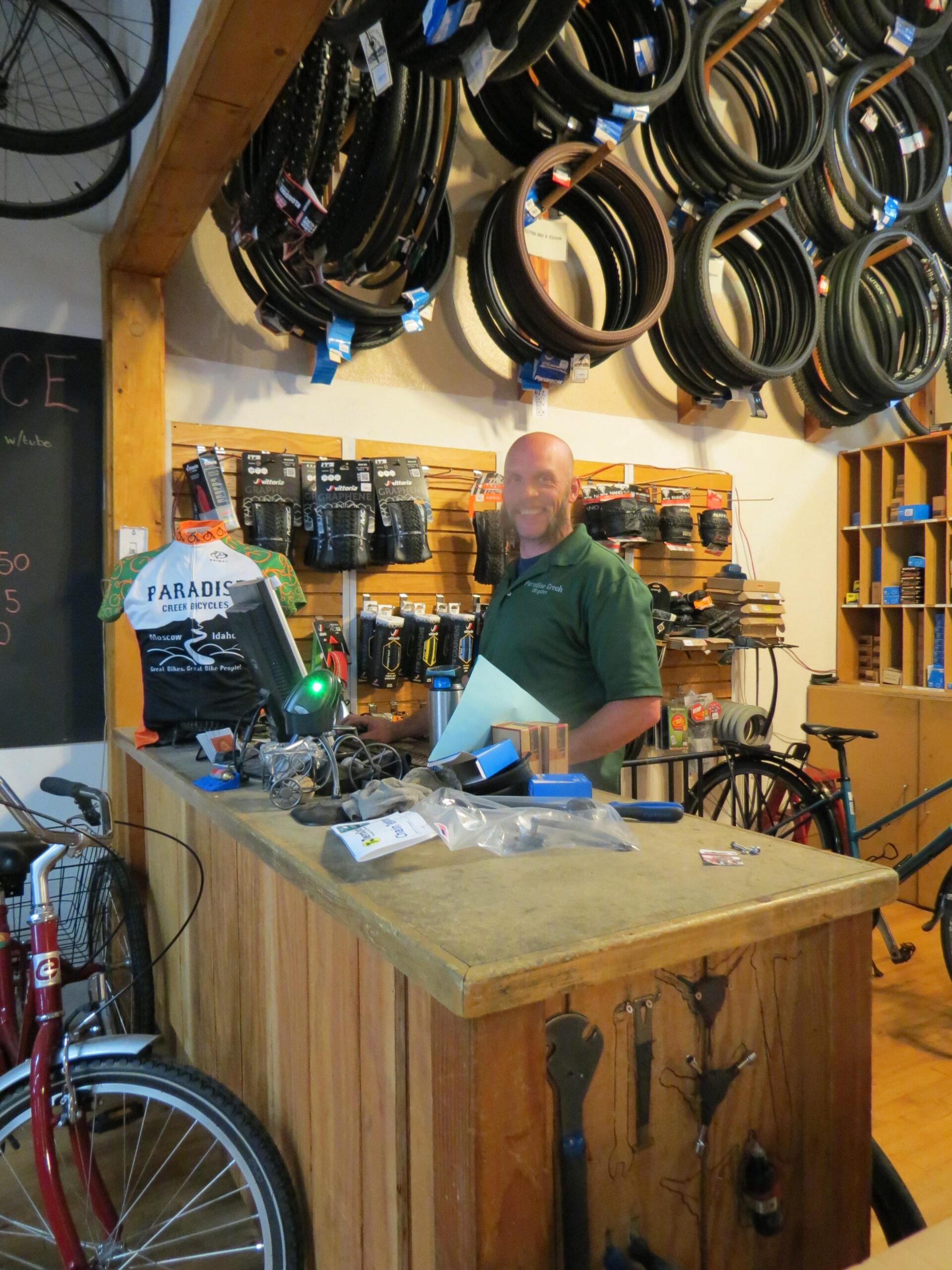 A bicycle shop interior featuring a smiling staff member standing behind a wooden counter. The walls are lined with various bicycle tires and accessories, while tools and equipment are out on the counter. A few bicycles are visible in the background.