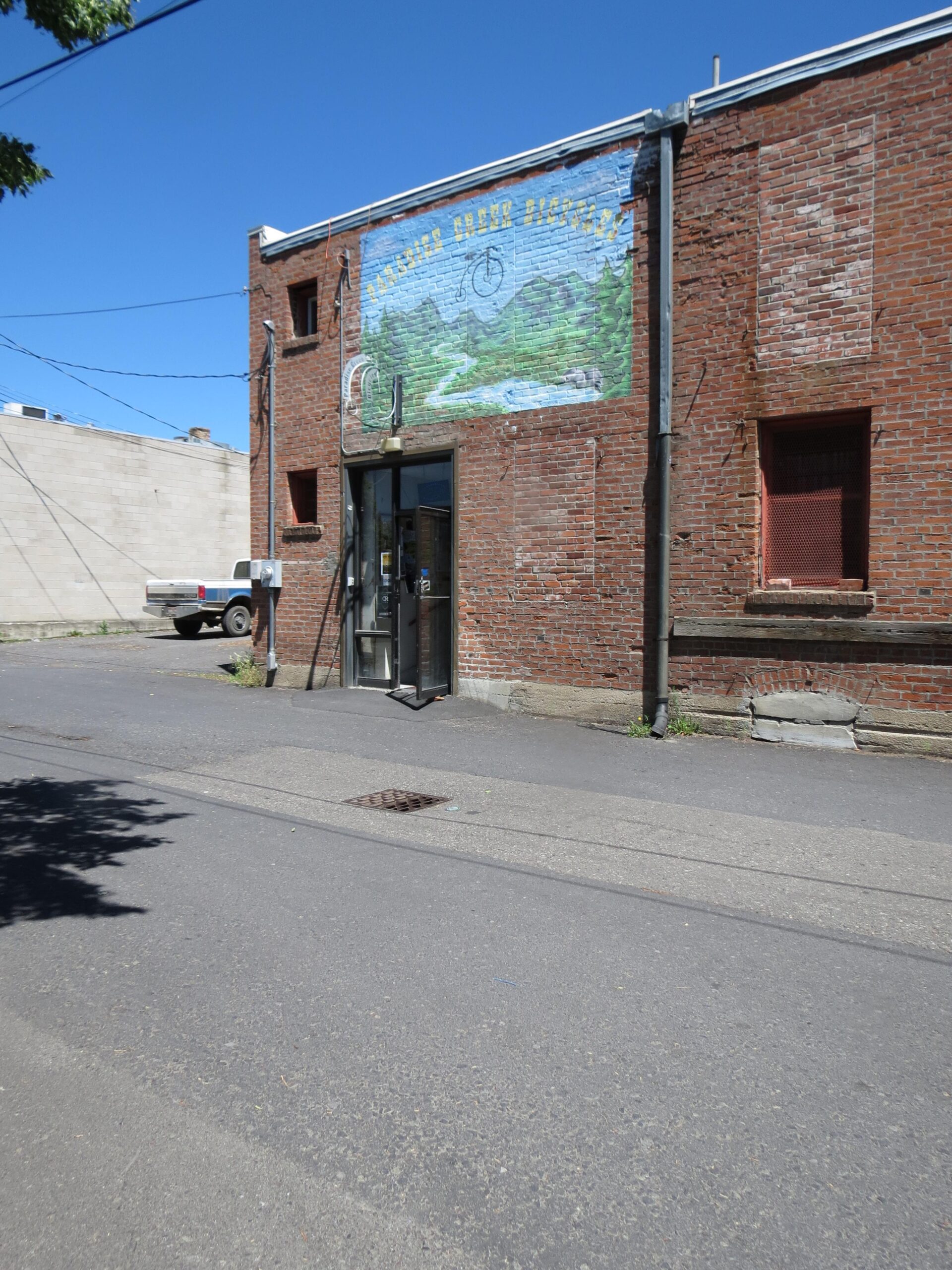 Alt text: A brick building with a colorful mural featuring a bicycle, mountains, and a river. The entrance has a glass door, and there are power lines overhead. A white truck is parked on the street nearby.