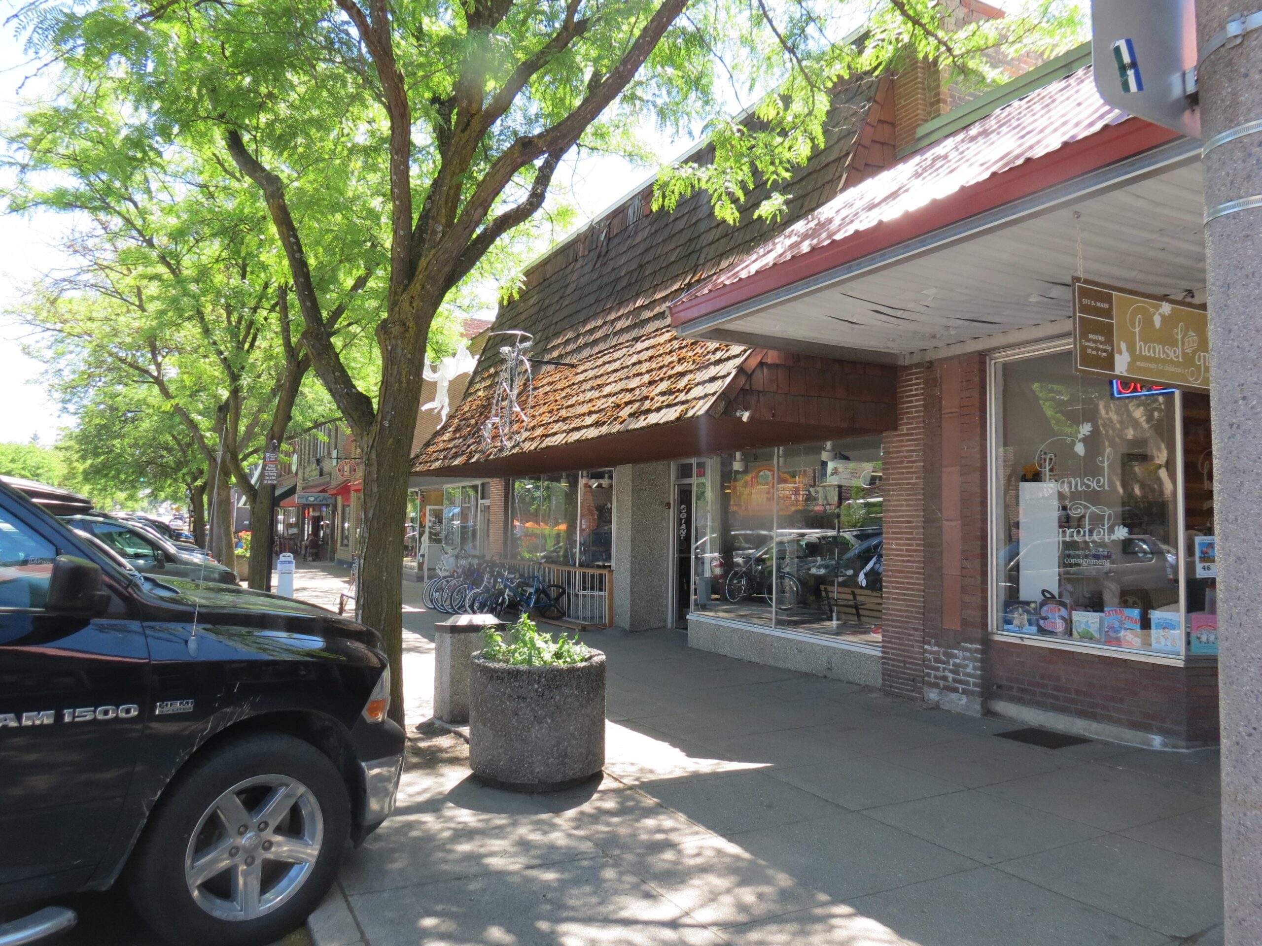 A sunny street scene featuring a retail store with large windows displaying products. The store has a sloped roof with shingles and a sign that reads "Hansel." Trees line the sidewalk, providing shade, and several parked cars are visible along the curb. Bikes are positioned outside the store, indicating a bike-friendly area.