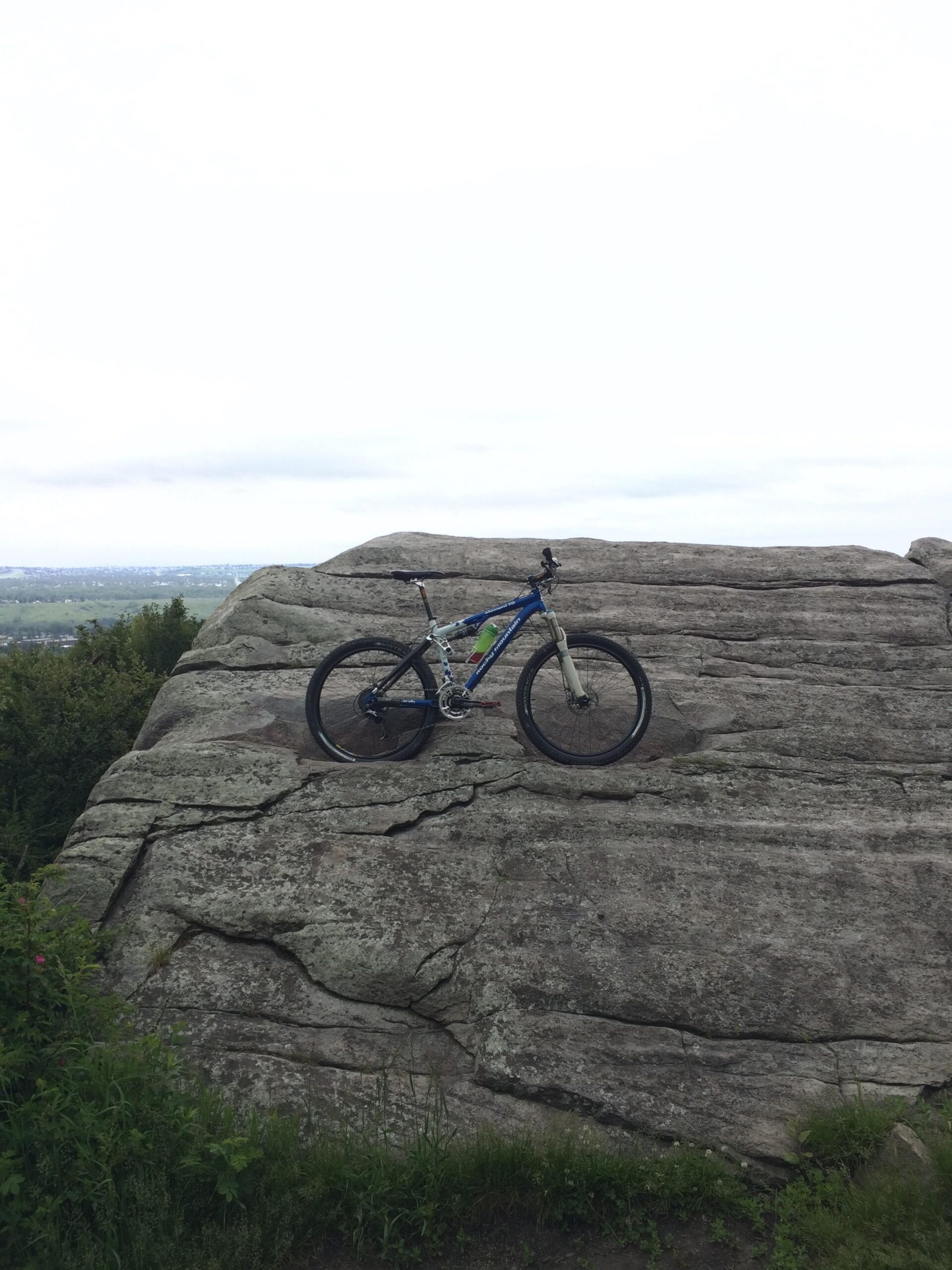 Rocky Mountain Element 70: A mountain bike resting on a large, flat rock formation, surrounded by greenery and a cloudy sky in the background.