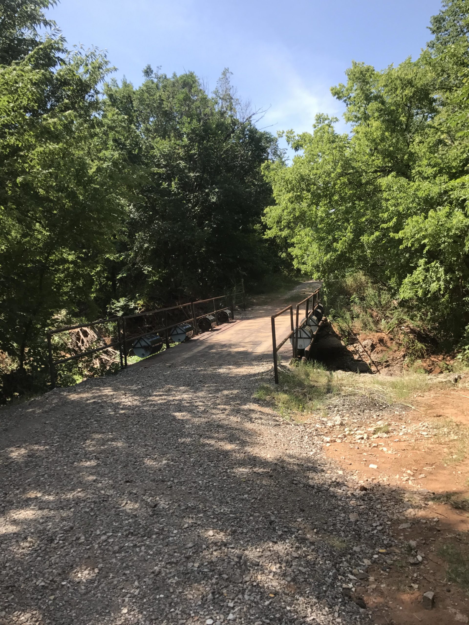 A narrow gravel path leading to a rustic metal bridge surrounded by lush green trees under a clear blue sky. The bridge spans over a creek, indicating a serene natural setting ideal for outdoor activities. SCIP Recreational Trail mountain bike trail.