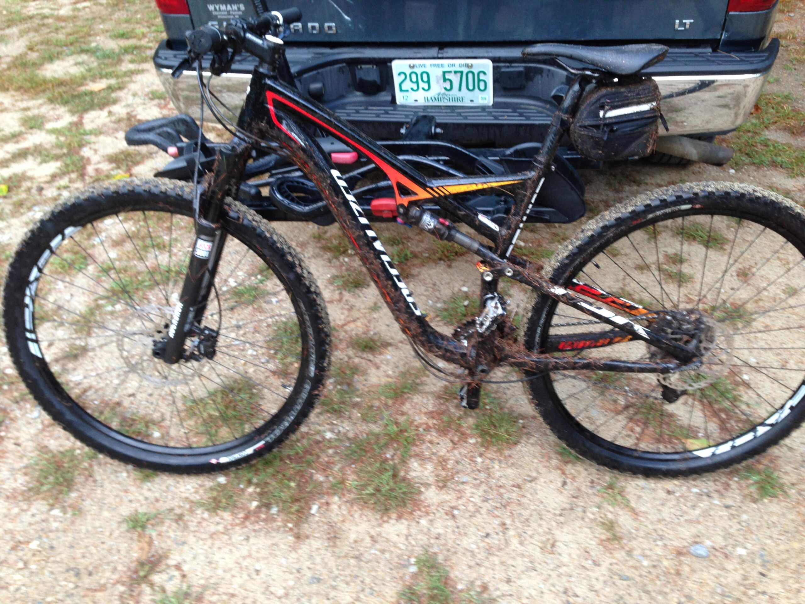 Specialized Camber Comp 29: A muddy mountain bike parked on gravel, with visible dirt splatters on the frame and wheels, alongside a pickup truck with a green license plate. The bike has a sleek design with black and orange accents.