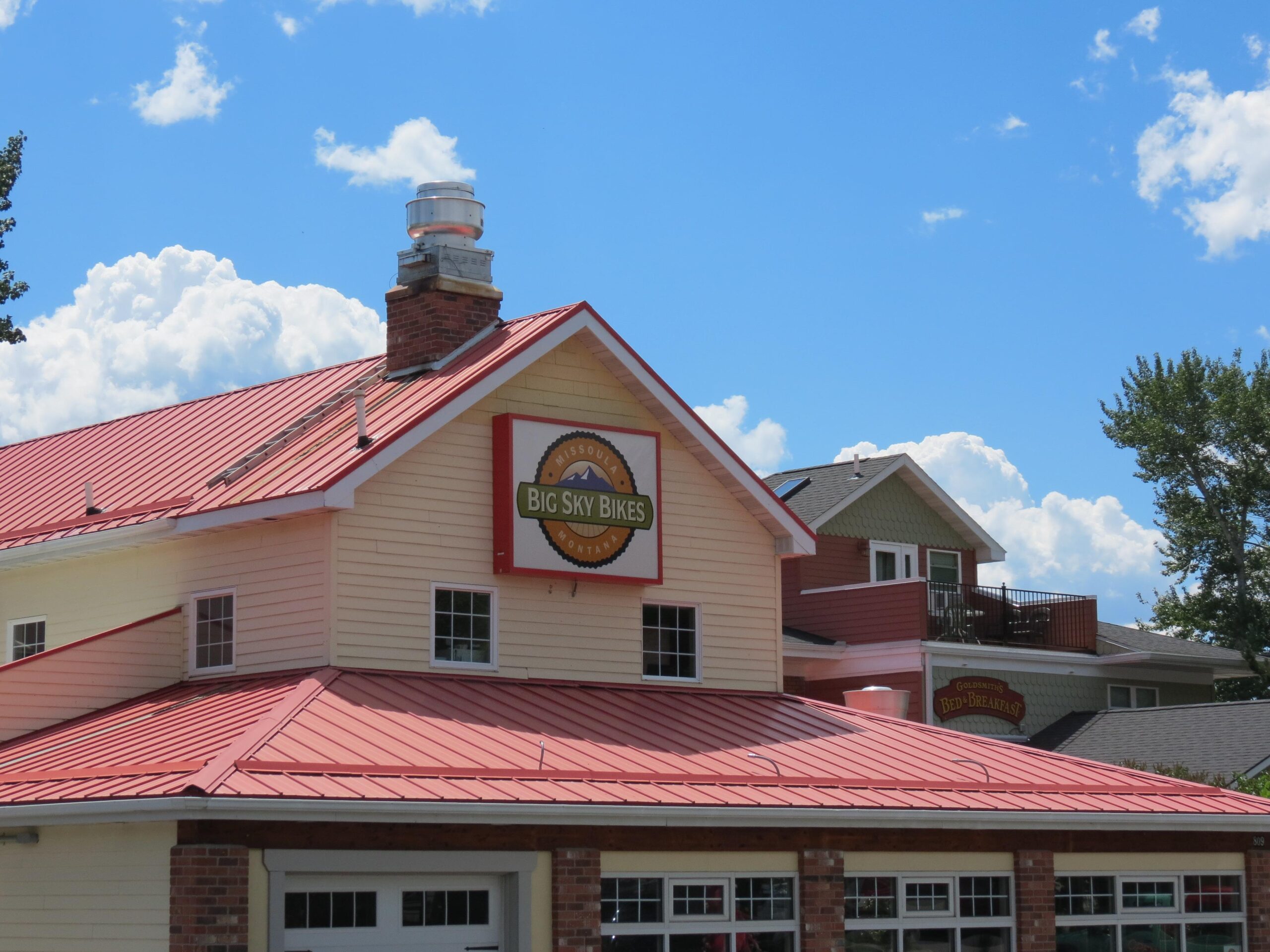 Building exterior with a red metal roof and a sign reading "Big Sky Bikes," located in a sunny setting with blue skies and white clouds. The building features yellow siding and multiple windows. Nearby, there are additional structures, including a green house with a balcony.