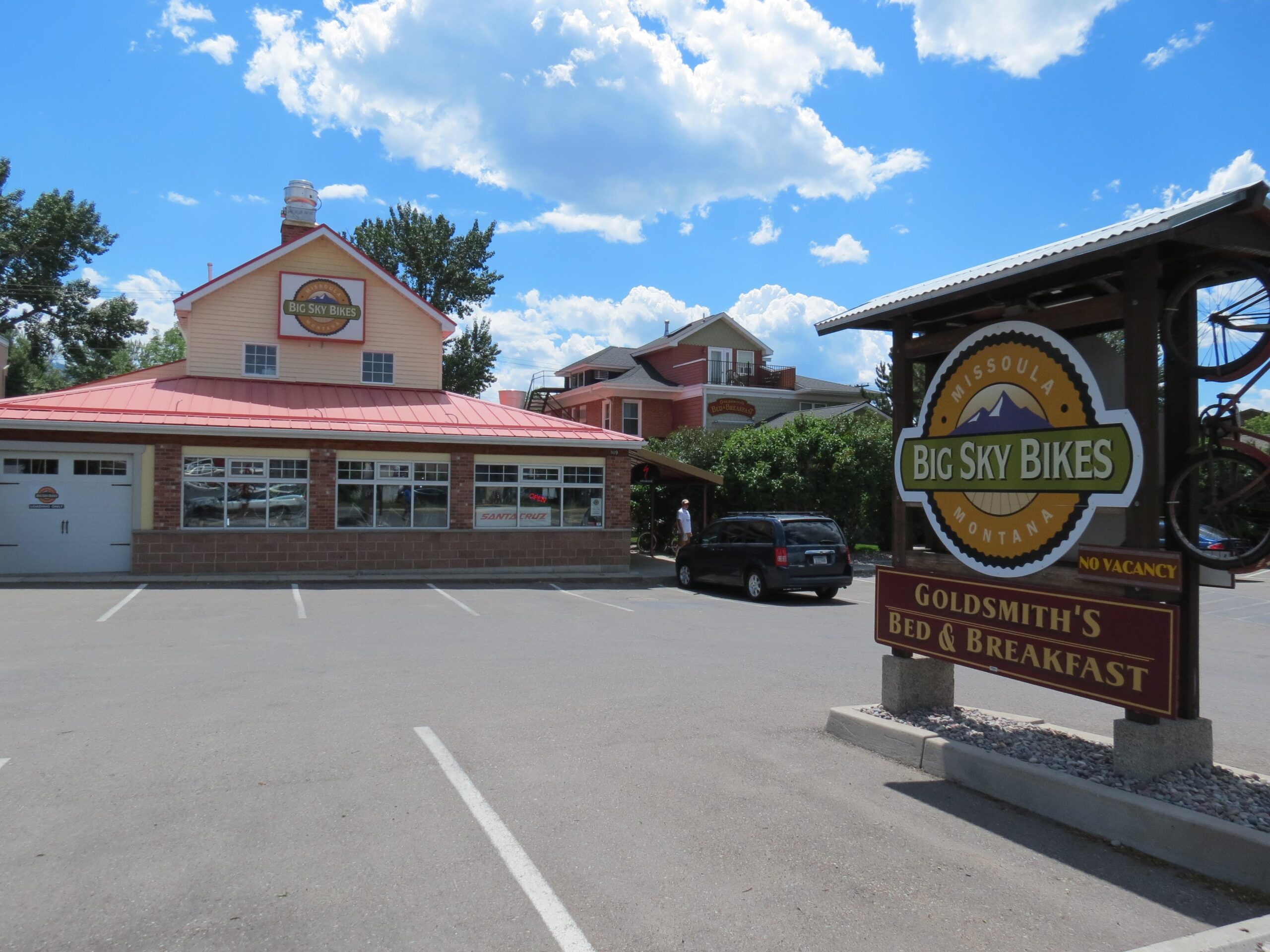 A bright summer day view of Big Sky Bikes, featuring a red-roofed building with large windows, located in Missoula, Montana. A sign for Goldsmith's Bed & Breakfast can be seen in the foreground, along with parked cars and clear blue skies.