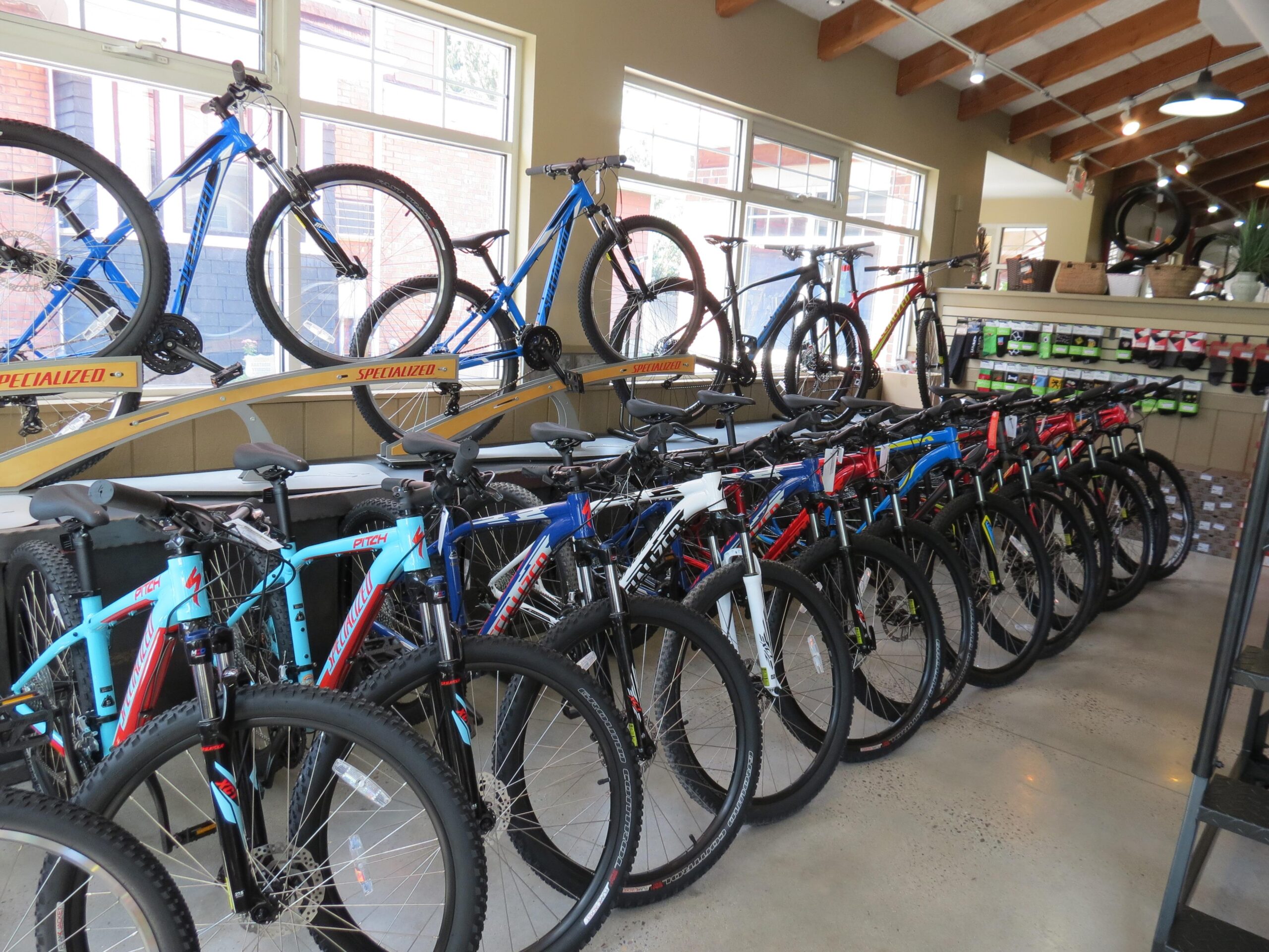 A display of various mountain bikes in a retail environment, showcasing a row of bikes in different colors, including blue and red, along with bikes mounted on racks in the background. The store features large windows allowing natural light to illuminate the space.