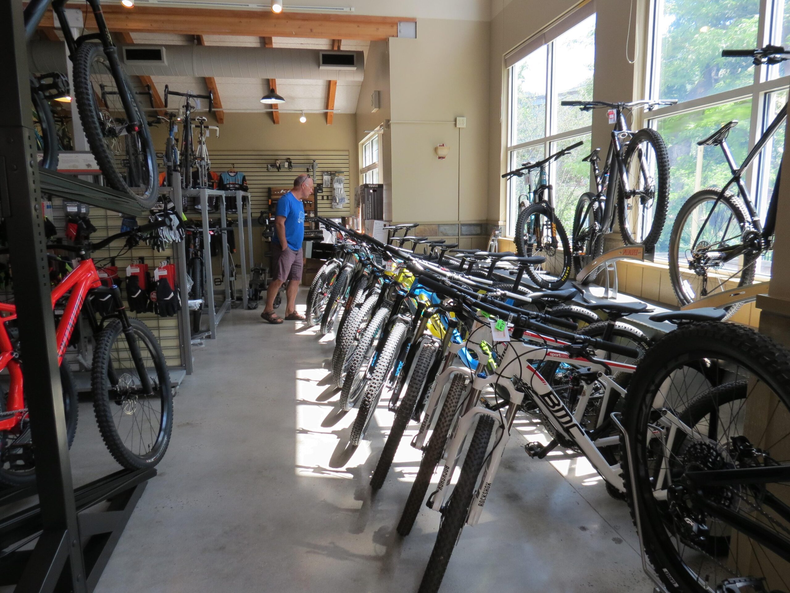 Interior of a bike shop displaying a variety of bicycles for sale. Several mountain bikes are lined up in the foreground, showcasing different colors and designs. A man wearing a blue shirt and shorts is engaged in examining the bikes. The shop has wooden beams and large windows that allow natural light to fill the space, creating a welcoming atmosphere.
