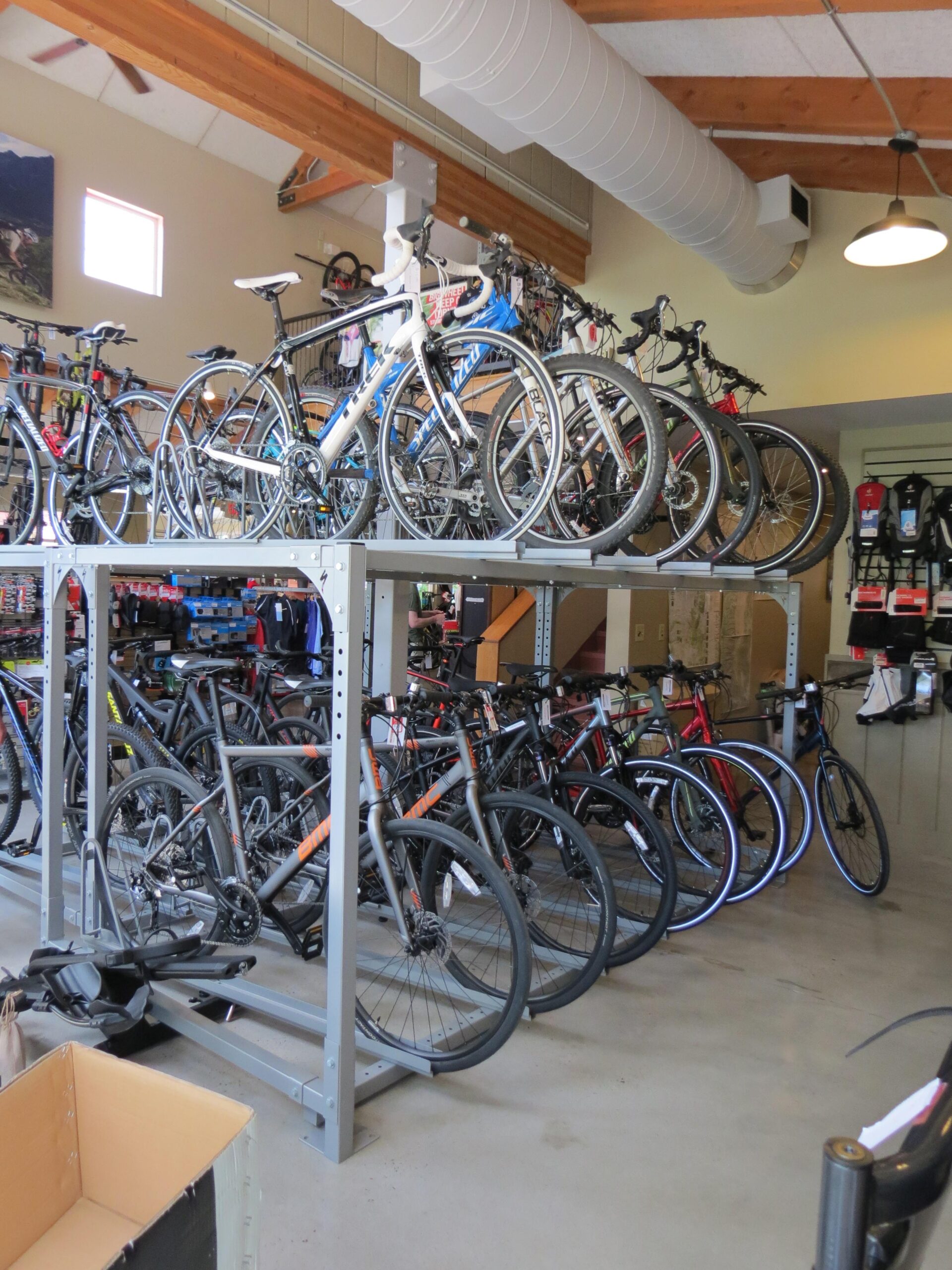 A bicycle shop interior showcasing various types of bicycles. Several bikes are displayed on a multi-level rack, while more bicycles are arranged on the floor below. The shop features a spacious and well-lit environment with wooden beams and shelves filled with cycling gear in the background.