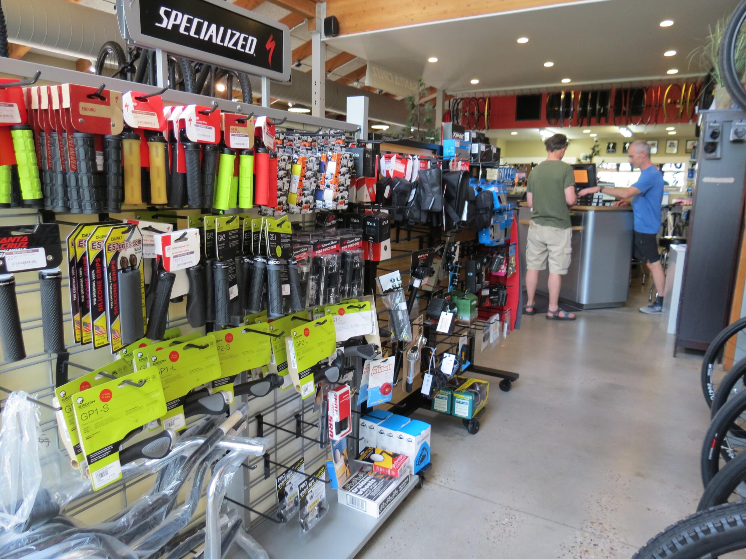 Interior of a bicycle shop featuring a variety of bike grips and accessories displayed on shelves. Two customers are interacting at the checkout counter, while bicycle parts and tools are visible throughout the store. The atmosphere is bright and inviting, showcasing a range of cycling equipment.
