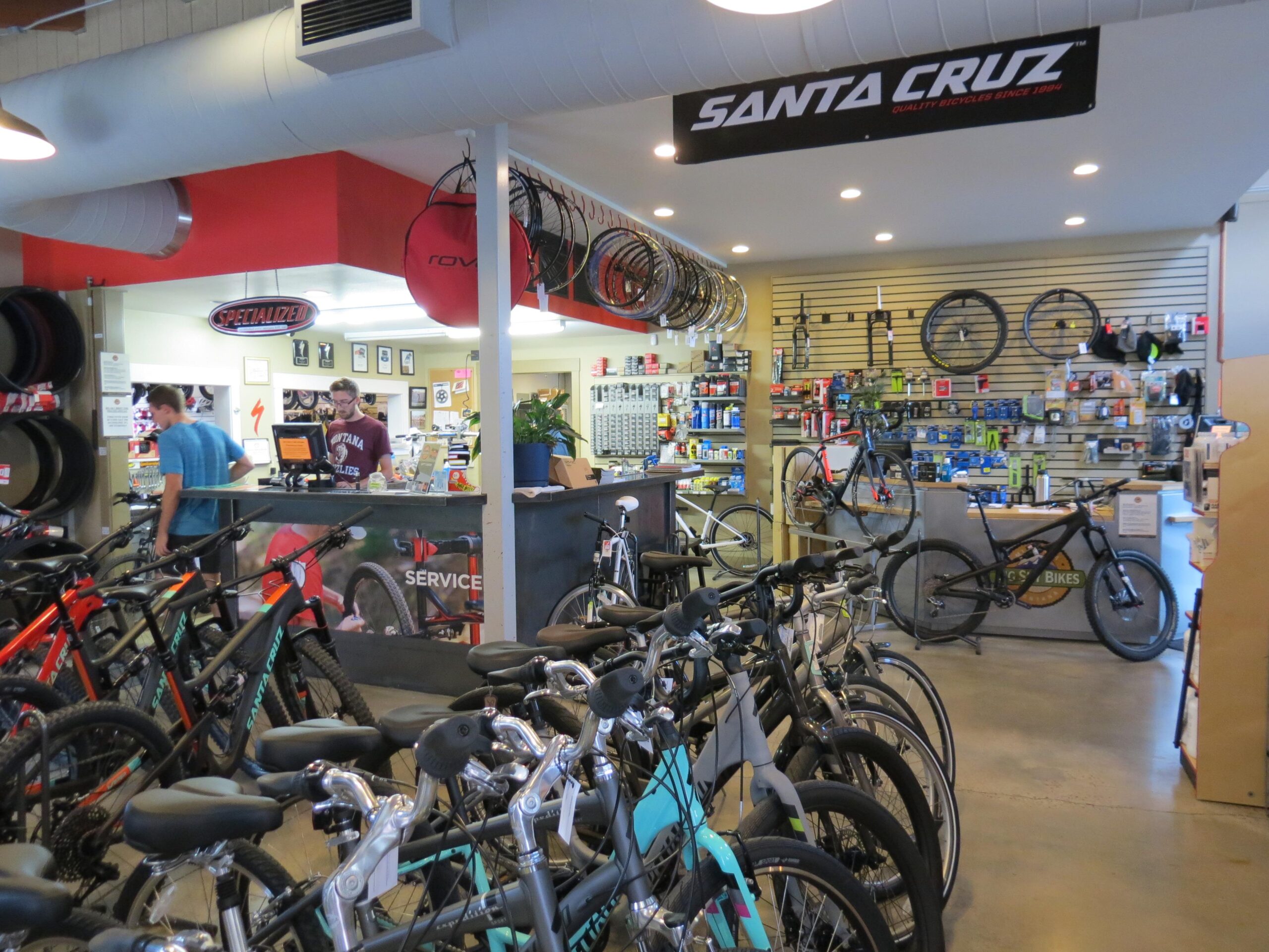 Interior view of a bicycle shop featuring various bikes for sale. The shop includes a service counter with staff assisting customers, and an array of bike accessories displayed on the walls. Signage for brands like Santa Cruz and Specialized is prominently visible. The space is bright and organized, showcasing a wide selection of mountain and road bikes.