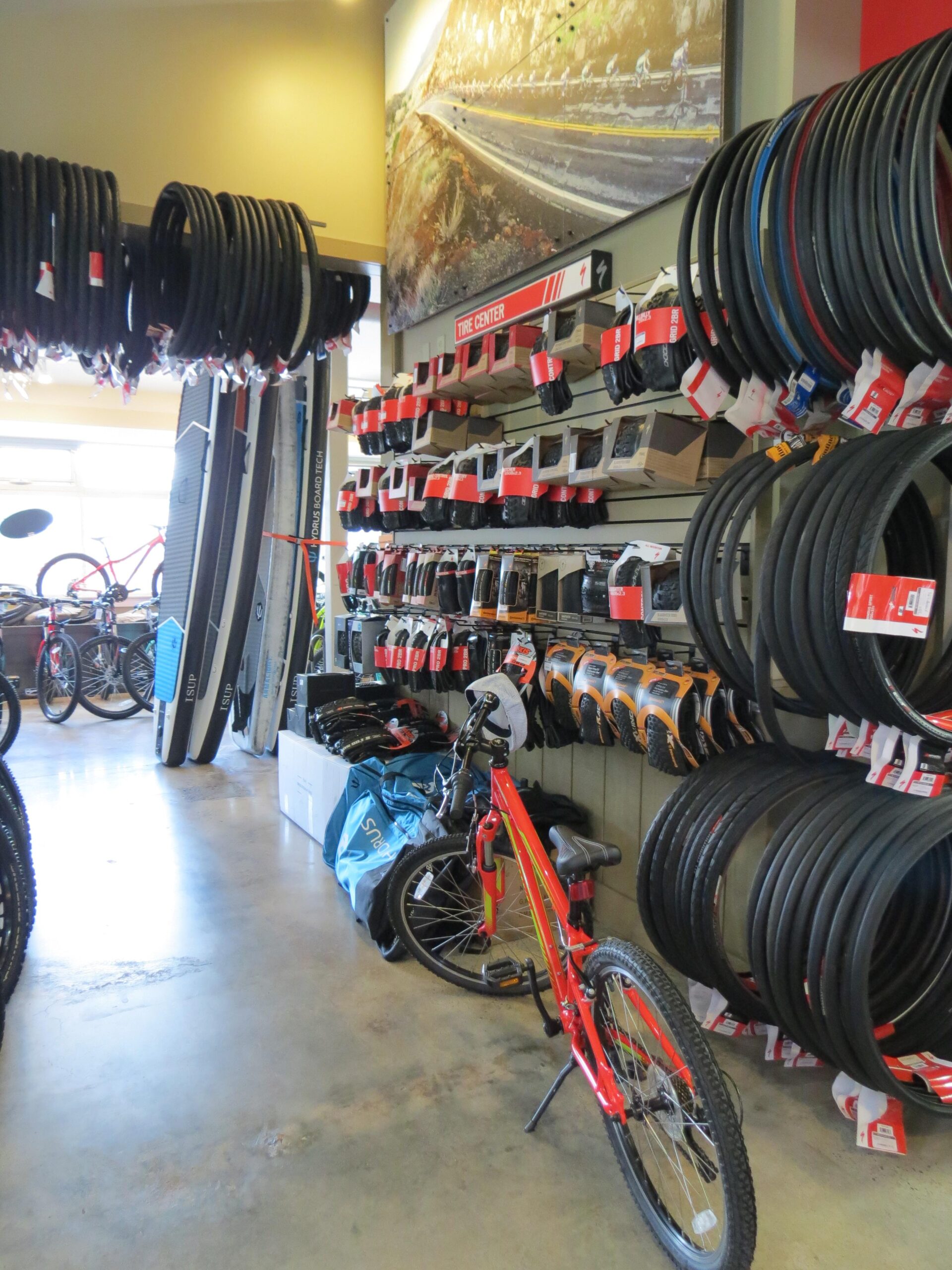 An interior view of a bike shop featuring a variety of bicycle tires displayed on shelves, along with a selection of bicycles. A red bike with yellow accents is positioned on the floor, and there are paddle boards leaning against the wall. The shop has a spacious layout with natural light coming in from large windows.